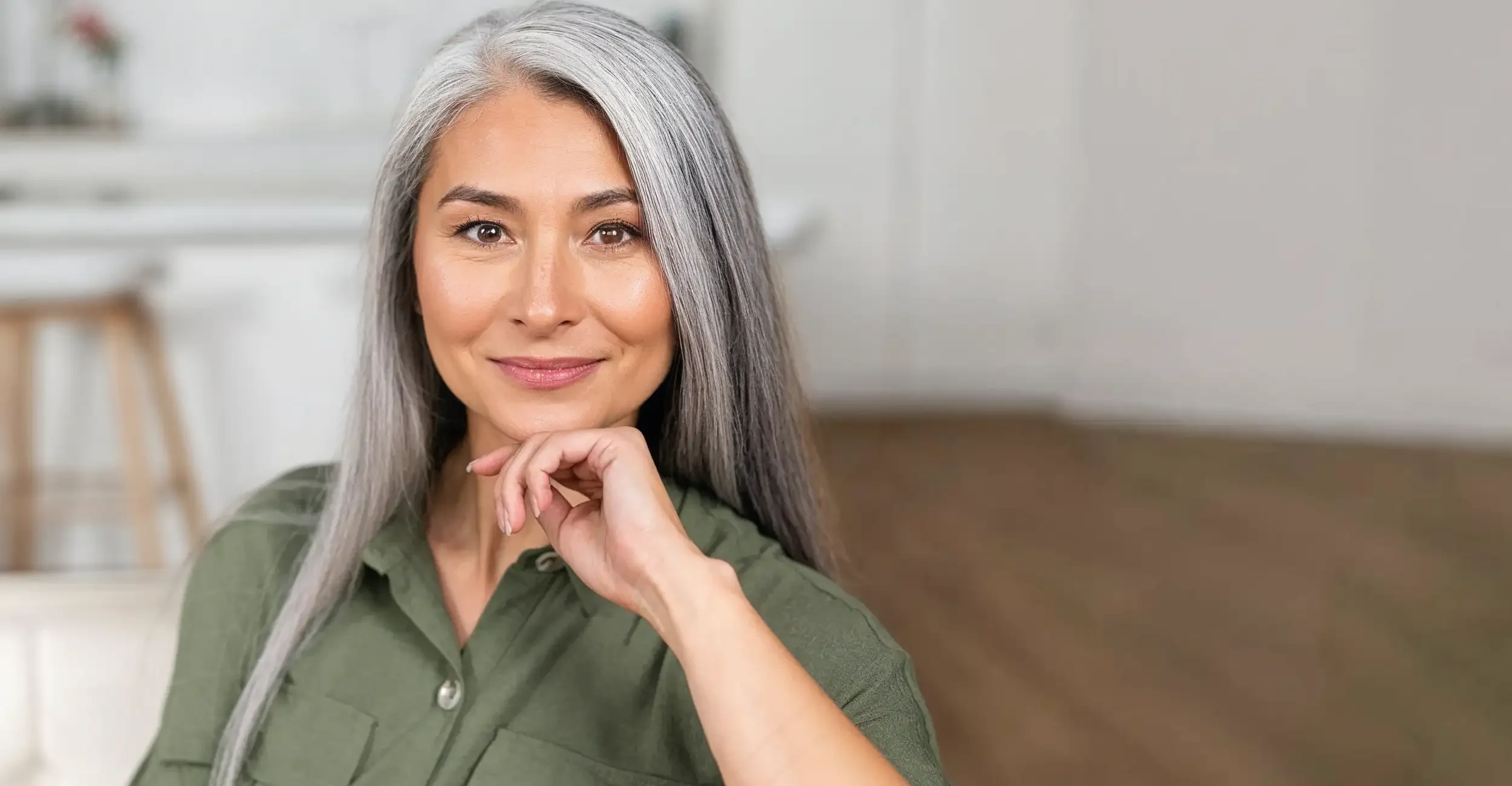 A woman with long gray hair, smiling softly, resting her chin on her hand, wearing a green shirt, in a bright indoor setting.