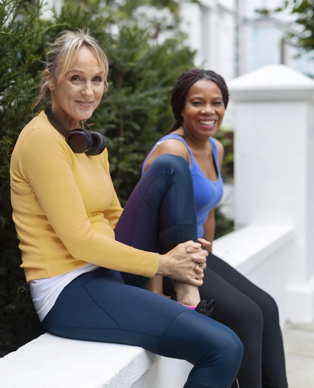 Two women sitting on a white wall outdoors, smiling. The woman on the left is wearing a yellow top and has headphones around her neck, while the woman on the right is wearing a blue top. Both women are dressed in athletic clothing and appear to be relaxing after a workout.