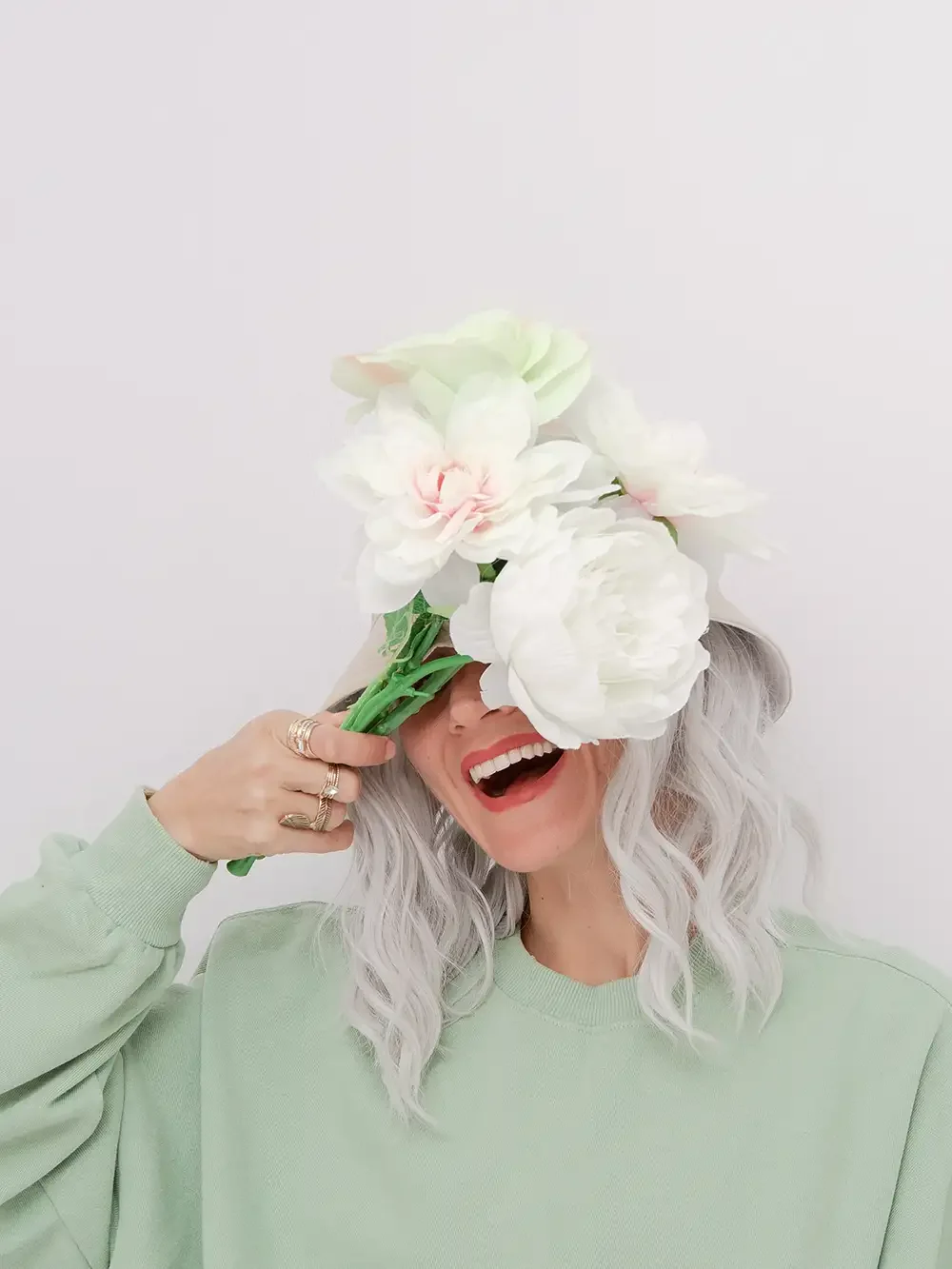 A woman with gray wavy hair, wearing a light green sweatshirt, holding a bouquet of large white and light pink flowers in front of her face, smiling and showing her teeth.