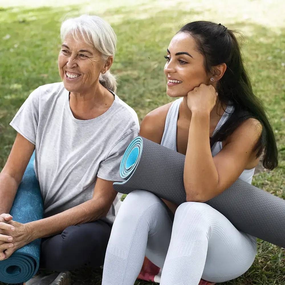 Two women, one elderly and one young, sitting on grass outdoors with yoga mats, smiling and enjoying each other's company.