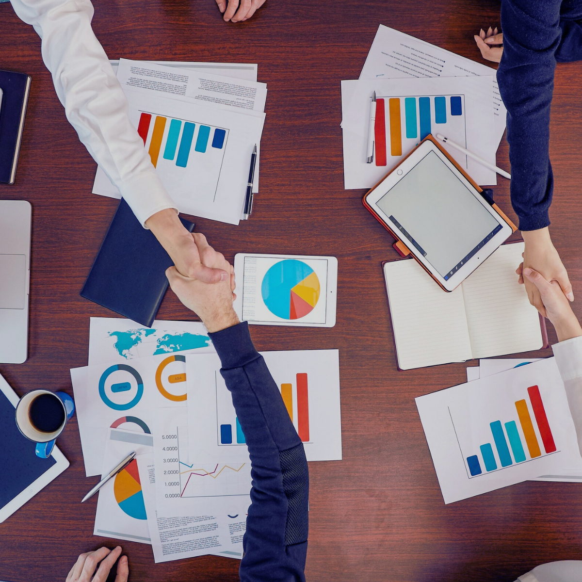 People shaking hands over a table filled with charts, graphs, notebooks, and digital devices in a business meeting.