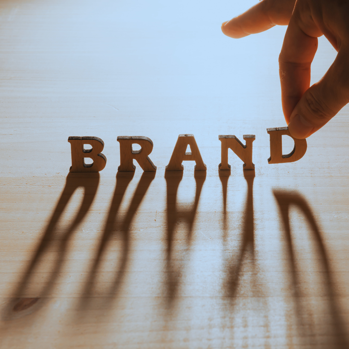 Person placing letter D on wooden blocks spelling 'BRAND' on a table, casting long shadows.