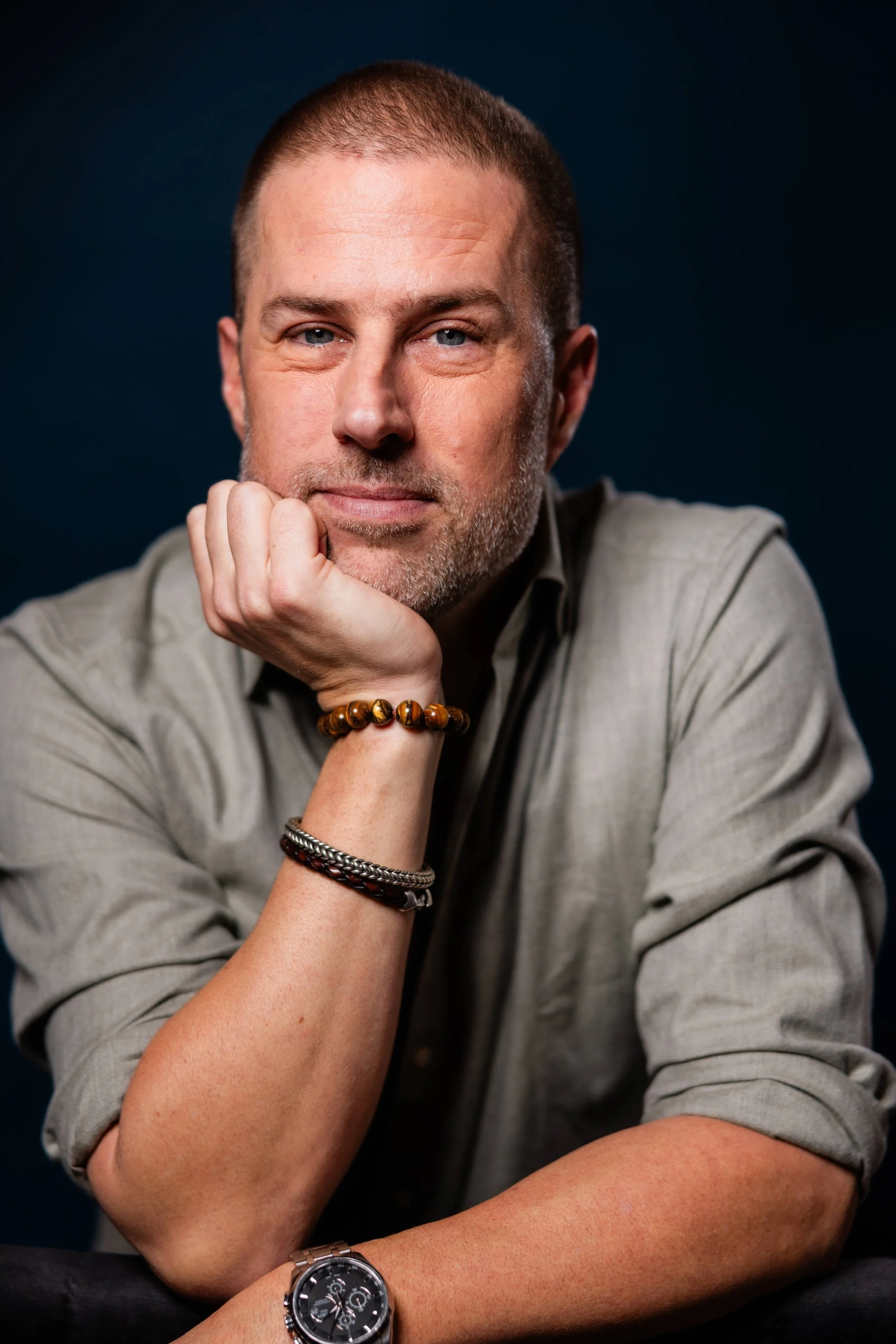 A middle-aged man with short hair and a beard, resting his chin on his hand, wearing a gray shirt, a black watch, and bracelets, looking directly at the camera against a dark backdrop.