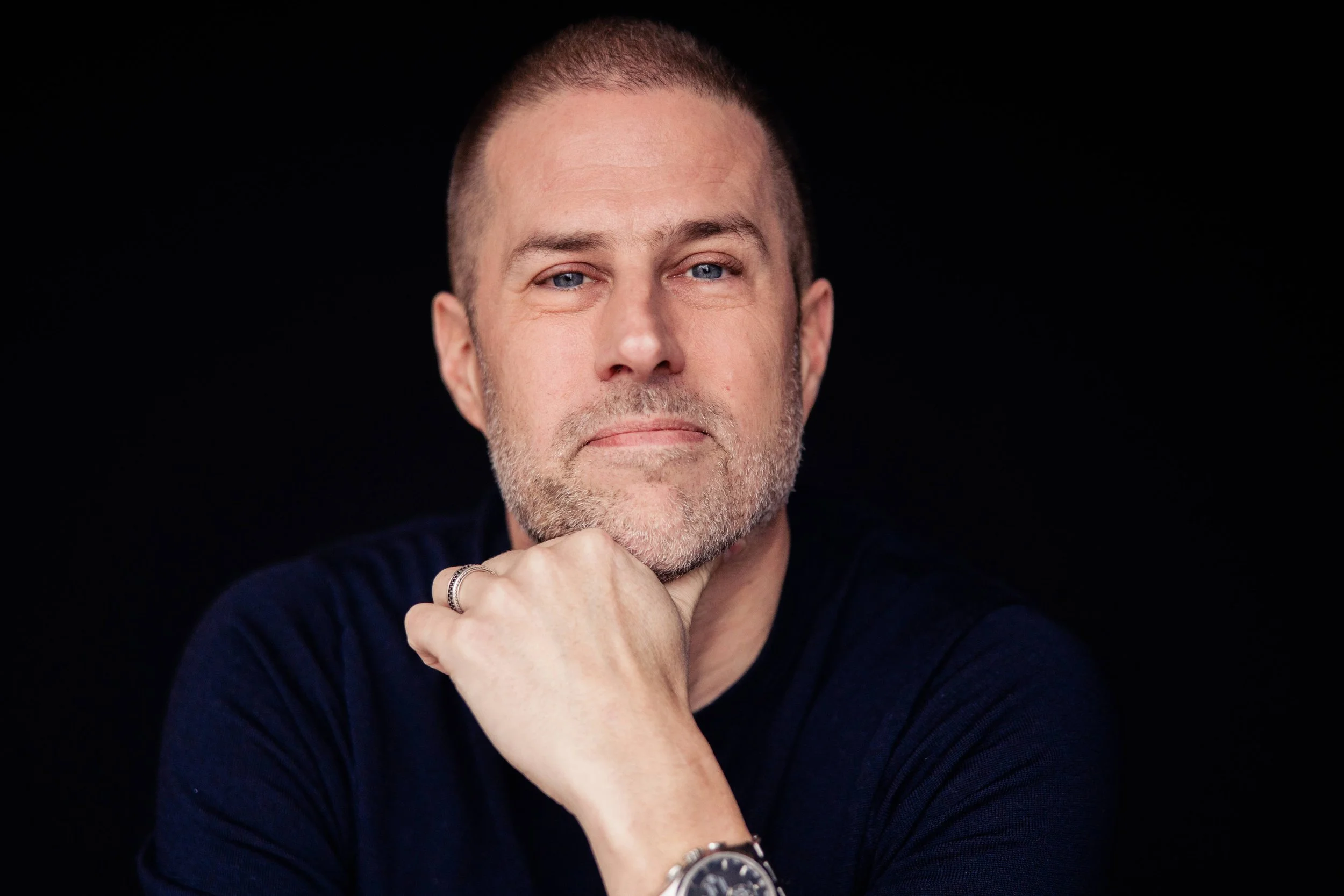 A man with short hair and a beard poses with his hand resting on his chin, wearing a dark shirt and a watch against a black background.
