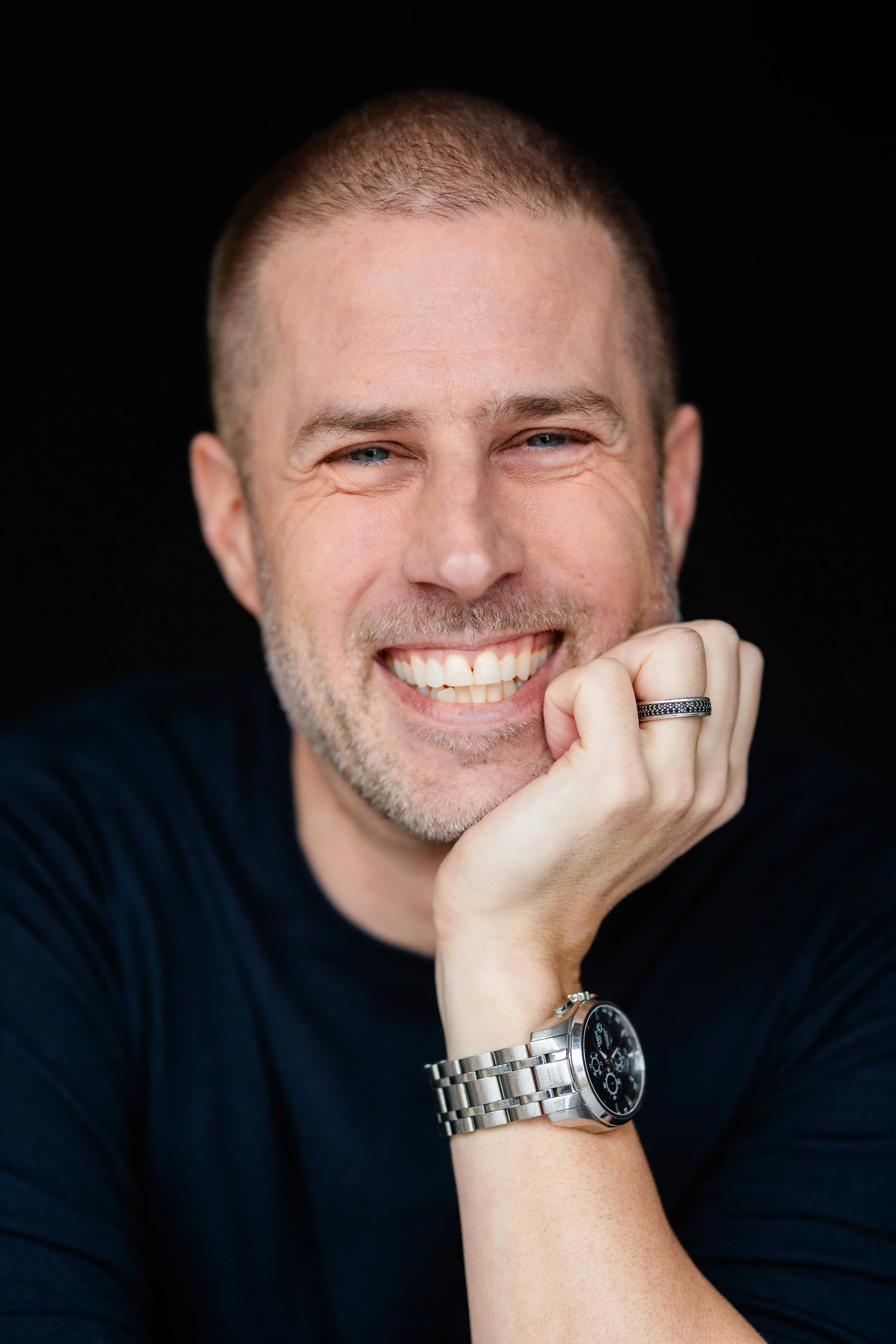 Close-up of a smiling man with short hair, wearing a black shirt, a silver watch, and a ring, resting his chin on his hand against a black background.