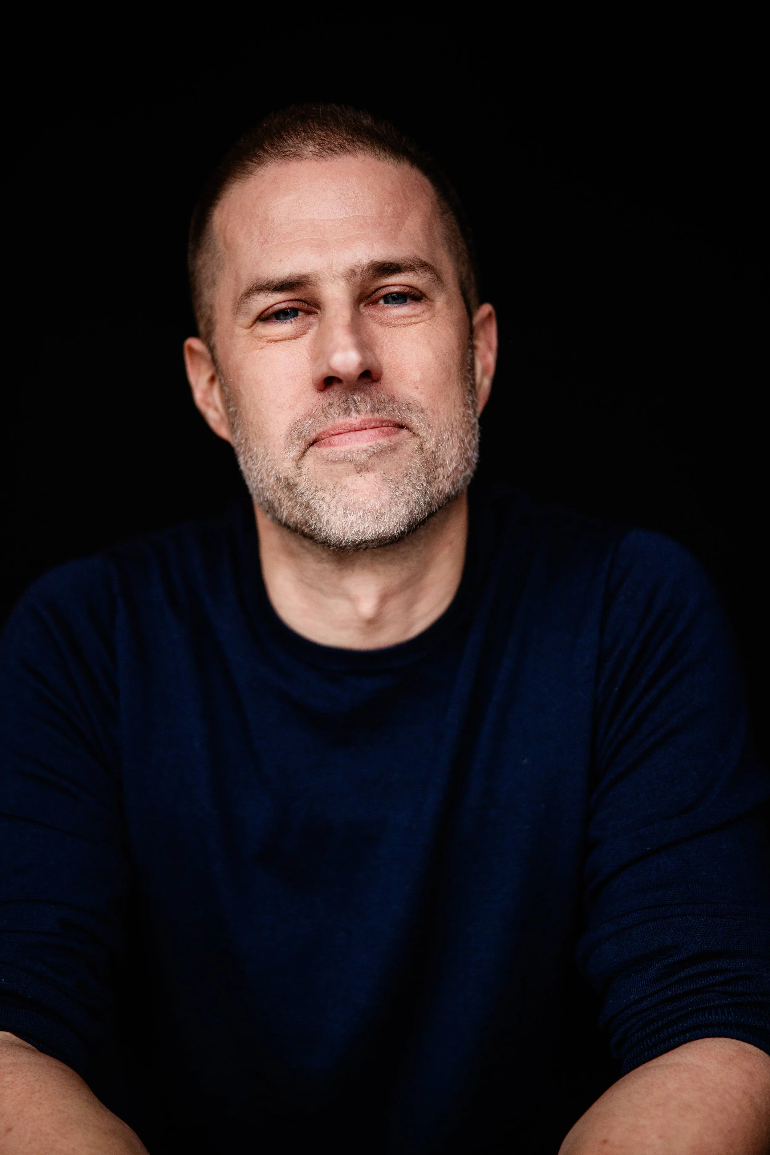 A close-up portrait of a middle-aged man with short brown hair, a beard, and blue eyes, wearing a navy blue shirt, against a black background.