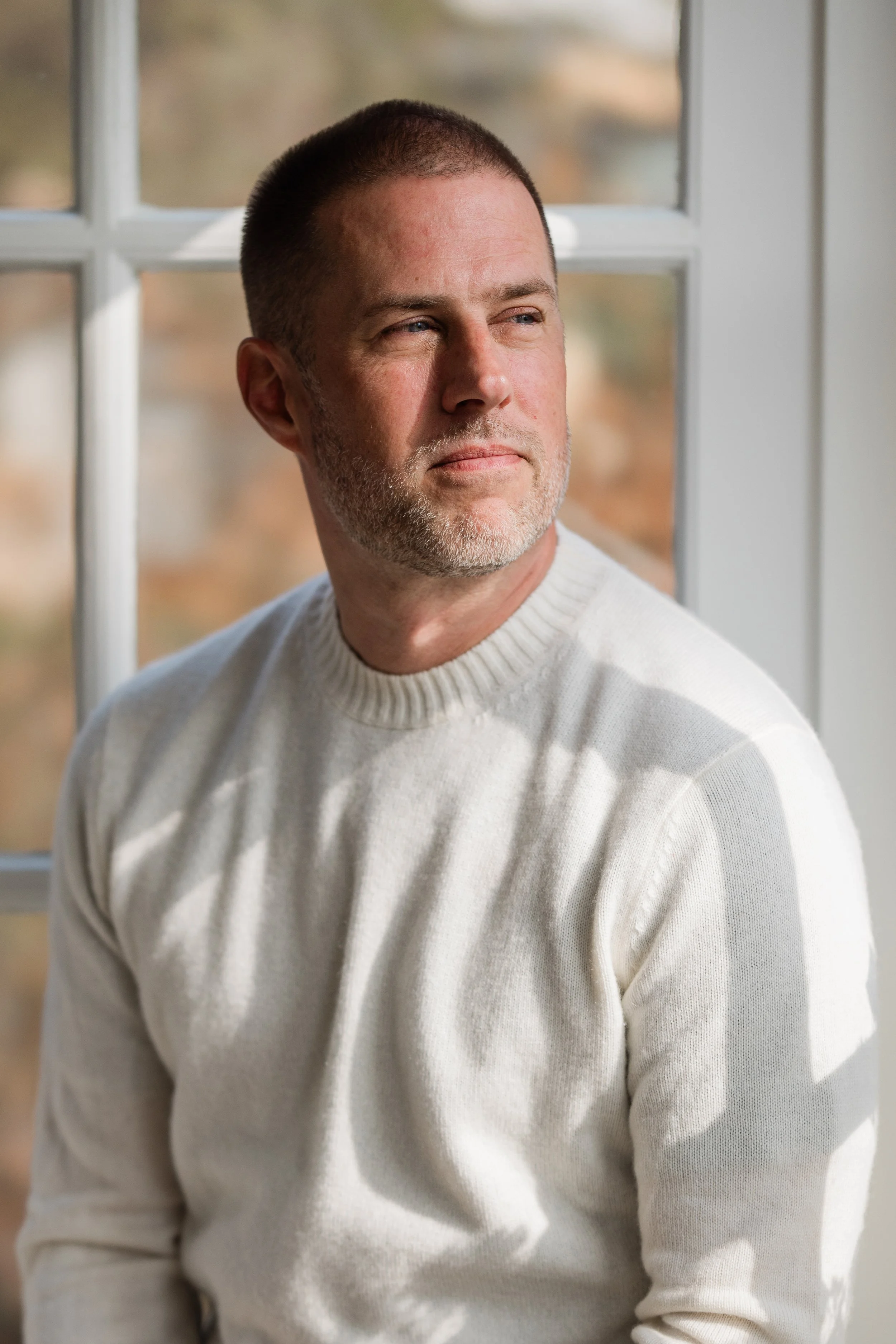 A man with short hair and a beard, wearing a cream-colored sweater, looking out a window.