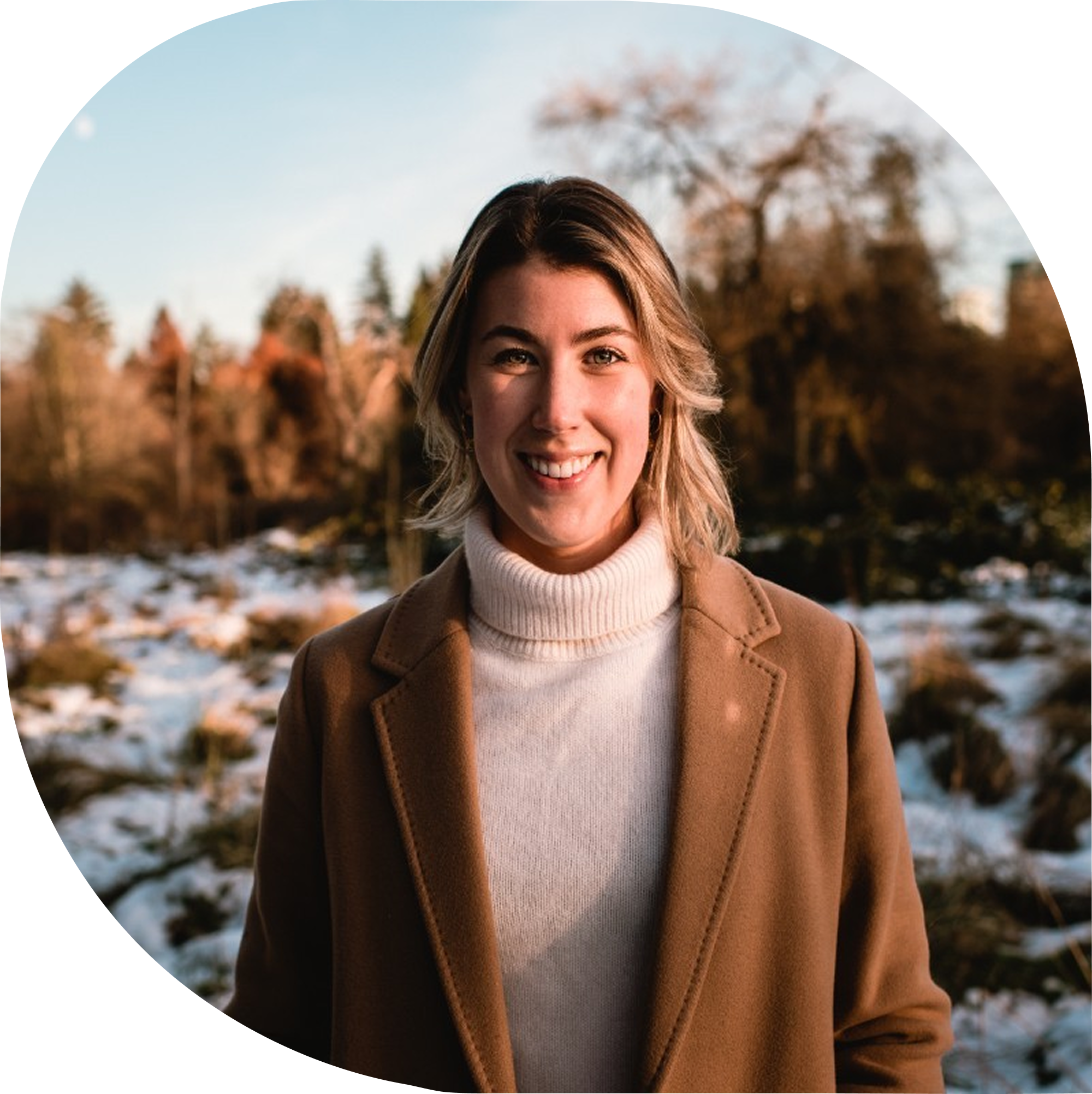 A smiling woman with shoulder-length blonde hair, wearing a tan coat over a white turtleneck, standing outdoors in a scenic area with snow, trees, and a clear sky in the background.