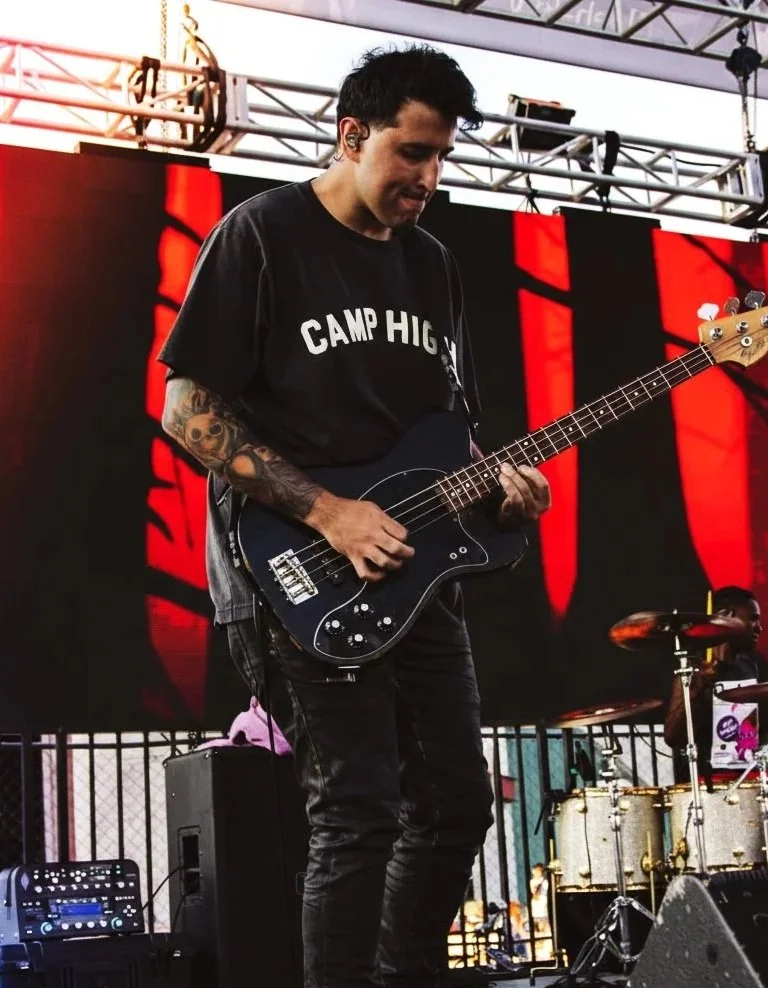 A musician playing a black electric bass guitar on stage, wearing a black t-shirt with 'CAMP HIGH' written on it, with tattoos on his arm, and standing in front of a large LED screen displaying red and black visuals.