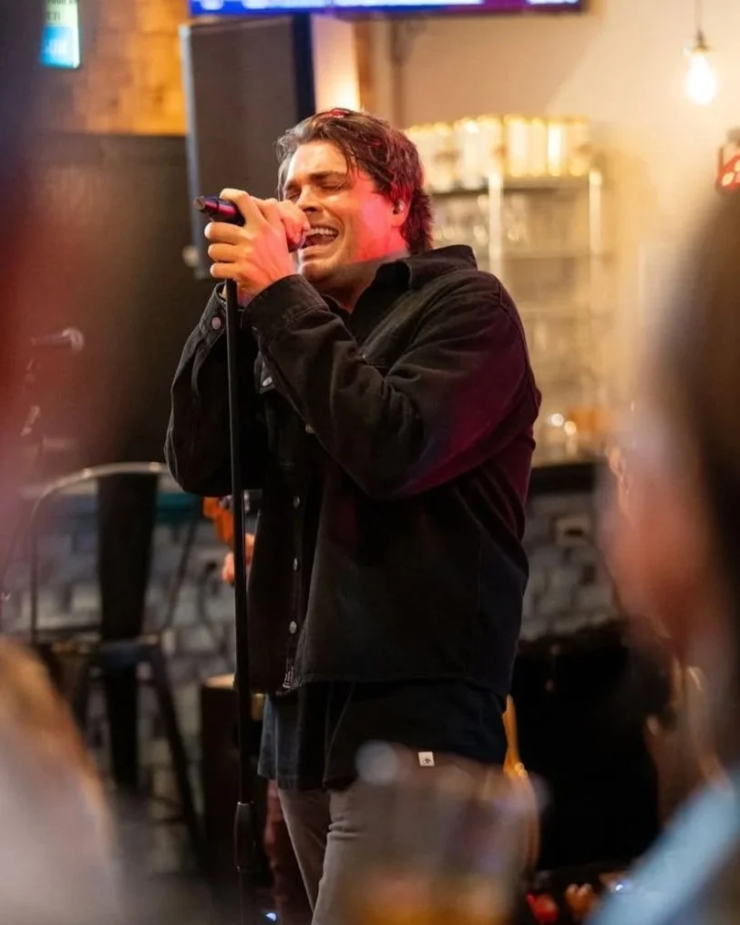 A man singing passionately into a microphone at a bar or restaurant. He has dark hair, is wearing a black jacket, and the background includes shelves with items and warm lighting.