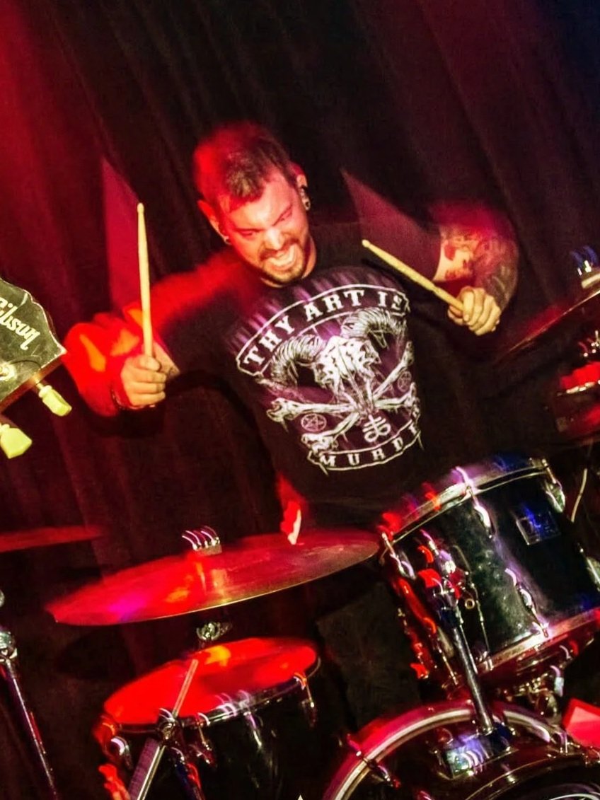 A drummer enthusiastically playing a drum set on stage, holding drumsticks and making an intense facial expression, with a dark background.