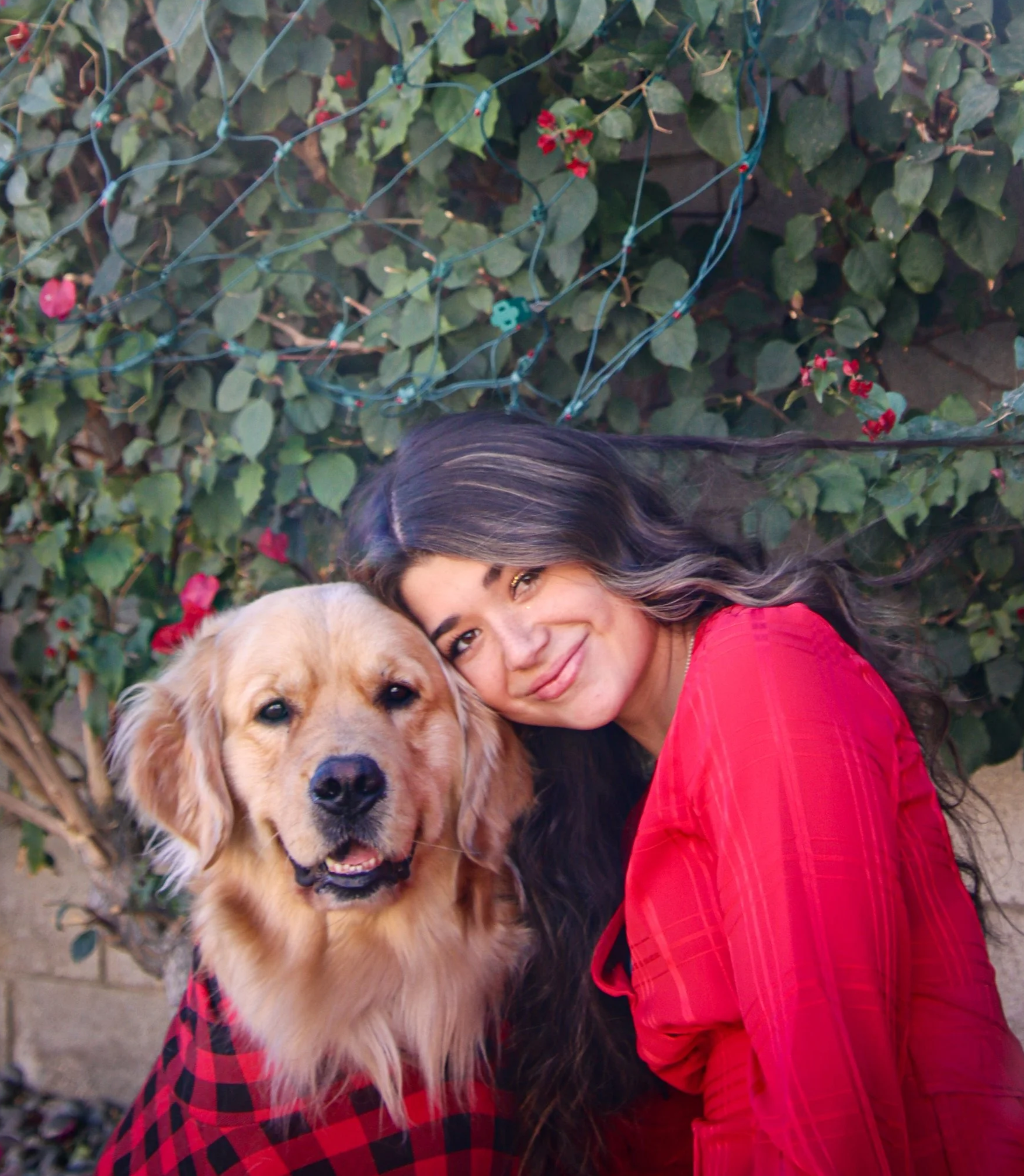 A woman with long dark hair and a golden retriever dog, both smiling, in front of a leafy background with string lights.