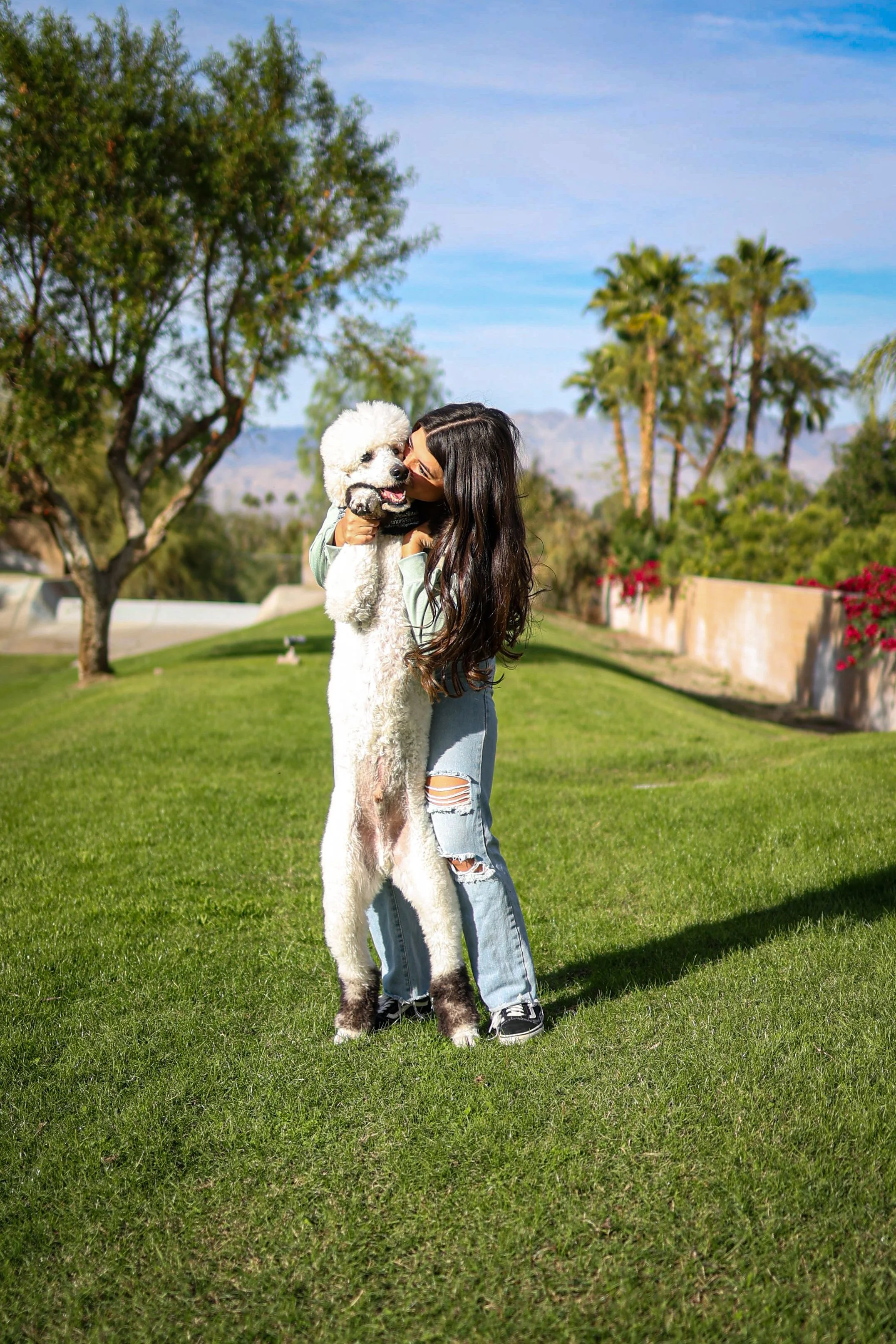 A woman with long dark hair hugging a large fluffy white dog outdoors on a sunny day.