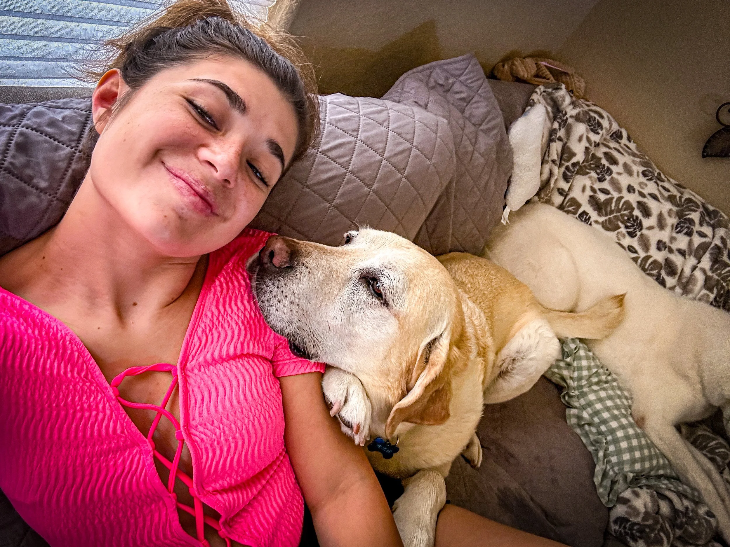 A woman with a pink top and a yellow Labrador Retriever lying on a bed, with the dog resting its head on her shoulder and appearing to give her a gentle kiss.