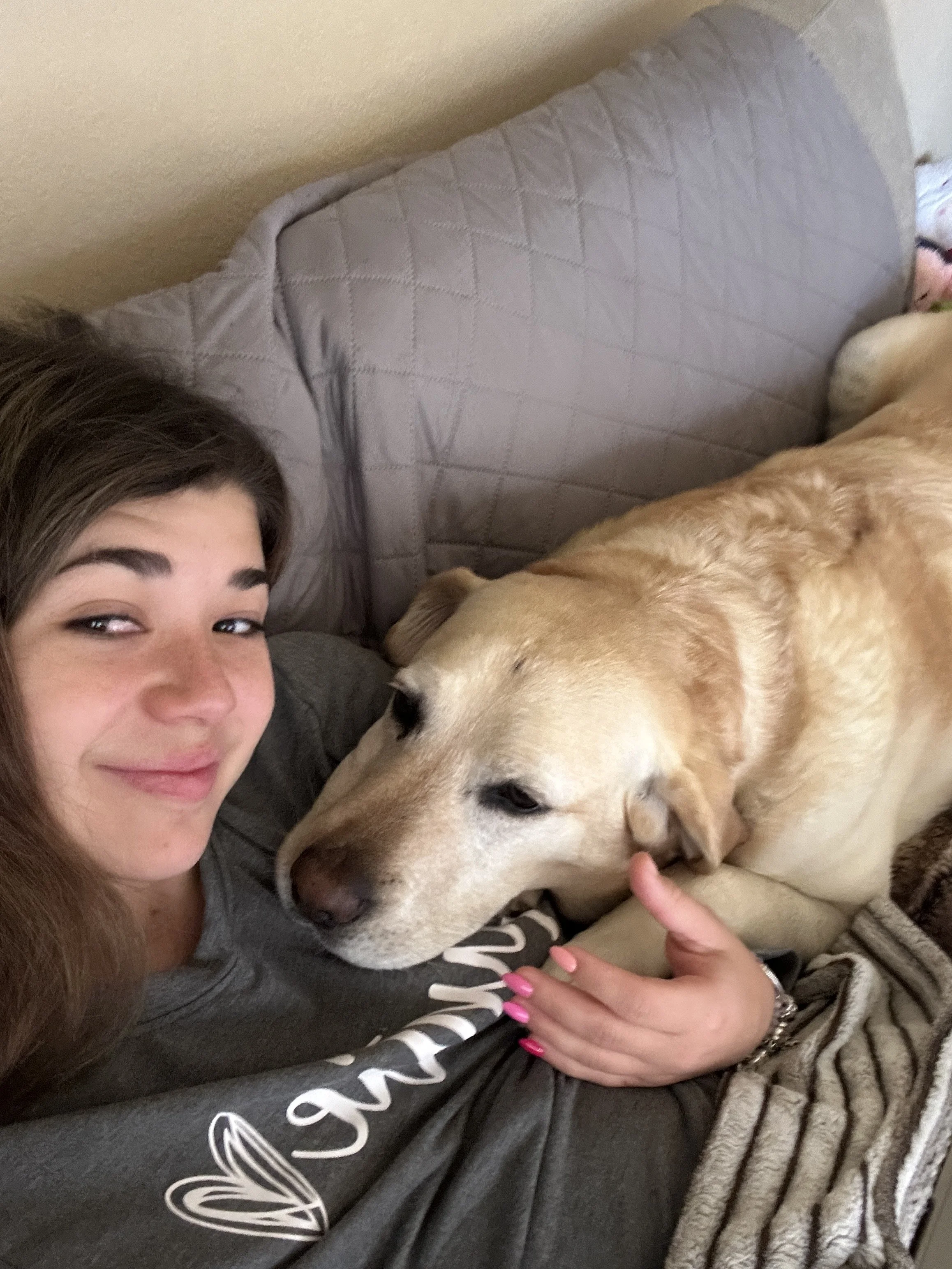 A woman with long brown hair lying on a couch next to a sleeping yellow Labrador retriever. The woman is smiling slightly, and the dog is resting its head on her chest-Dog Board & Train in La Quinta CA