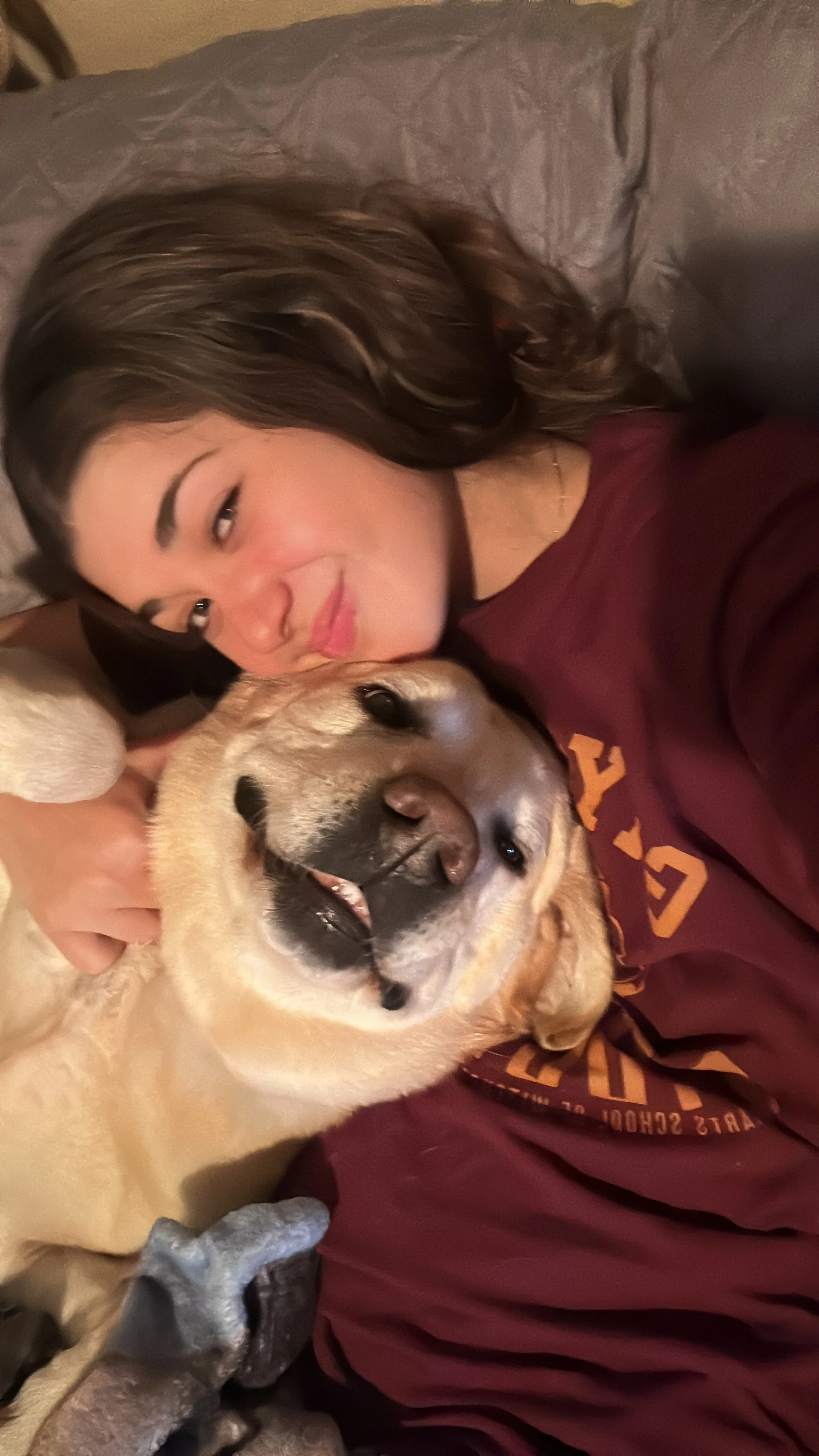 A young woman with long wavy hair and a maroon sweatshirt lying on a bed, smiling with her eyes closed, cuddling a large, smiling yellow Labrador retriever with a black nose-Dog Board & Train in La Quinta CA