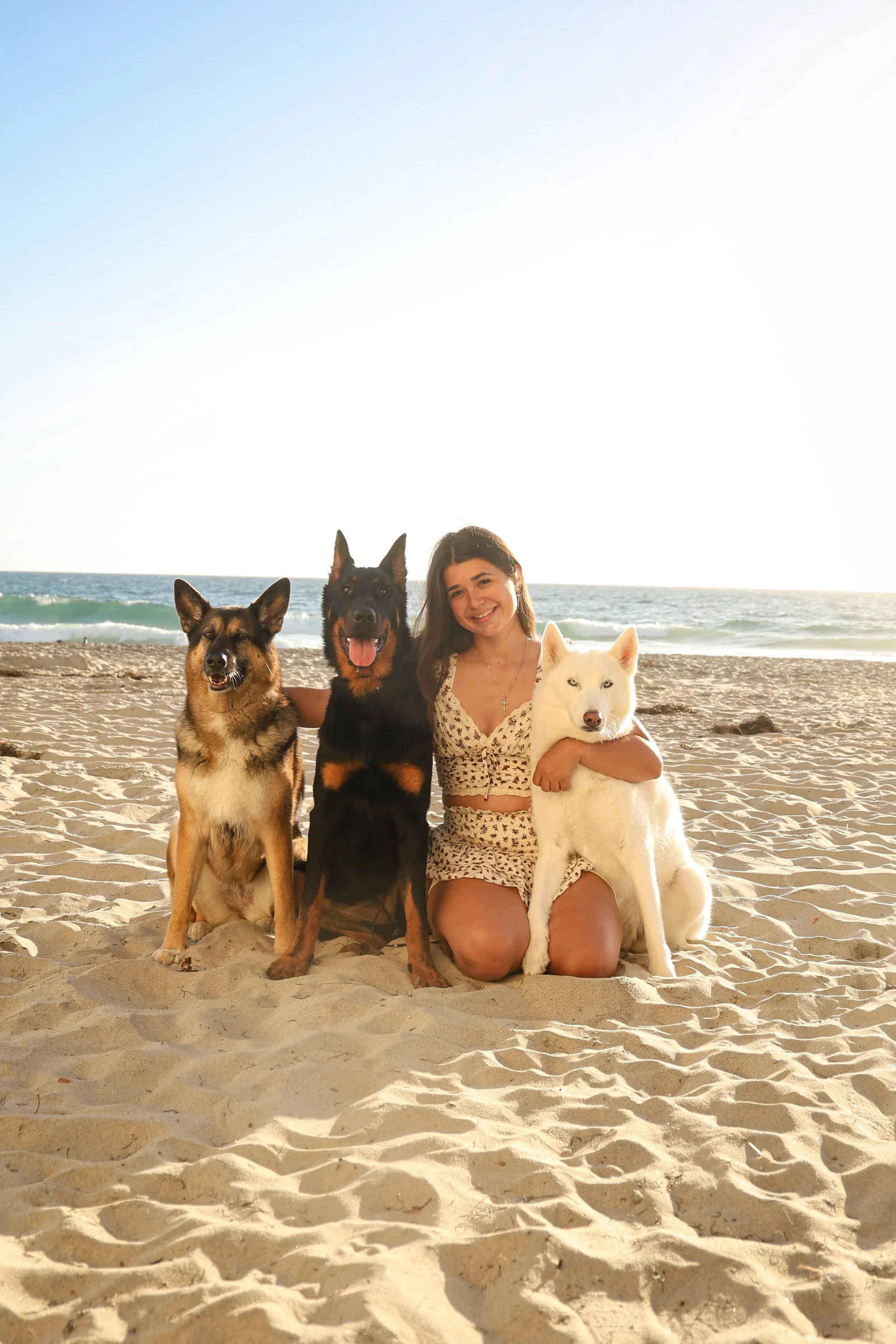 A woman kneeling on the beach with three dogs, smiling, with ocean waves and a clear sky in the background.