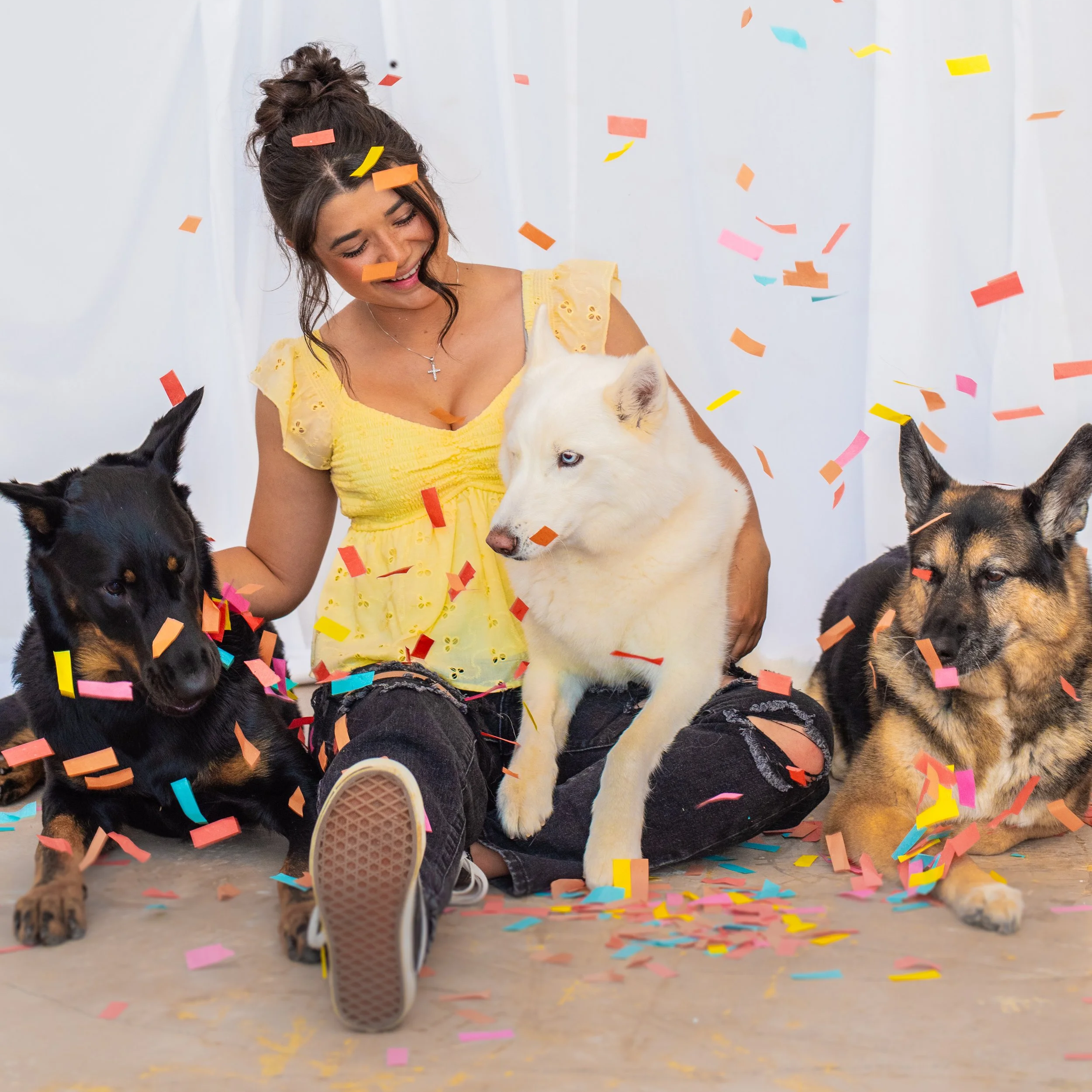 A young woman with dark hair in a bun, wearing a yellow dress, is sitting on the floor surrounded by falling colorful confetti and puppies, smiling with her eyes closed.