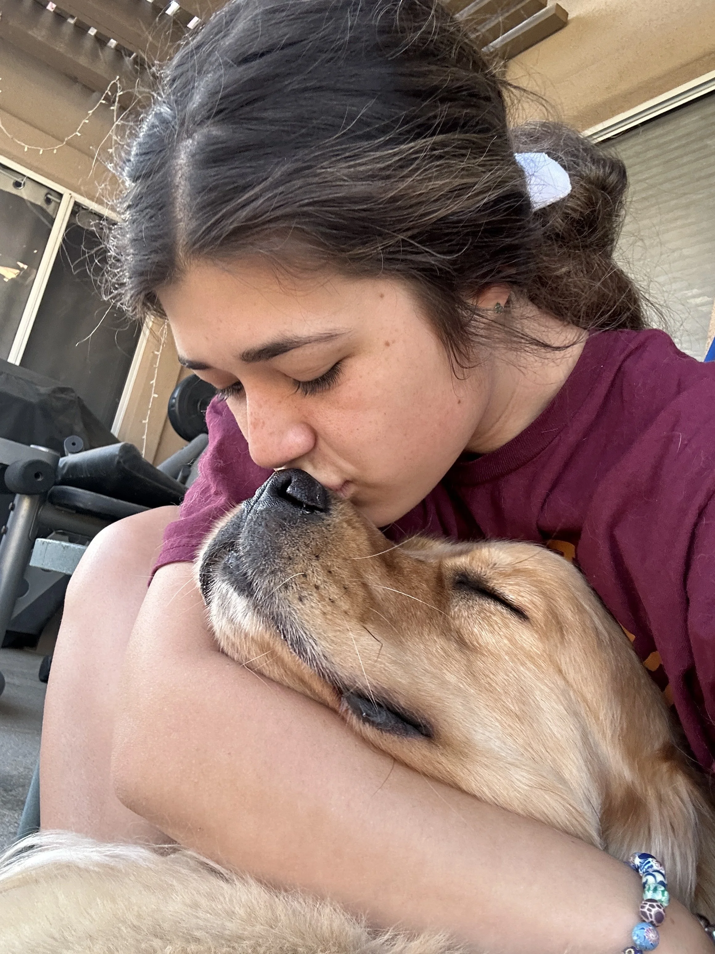 A young woman with dark hair and freckles is kissing a sleeping golden retriever dog on the nose while cuddling it -Dog Board & Train in La Quinta CA