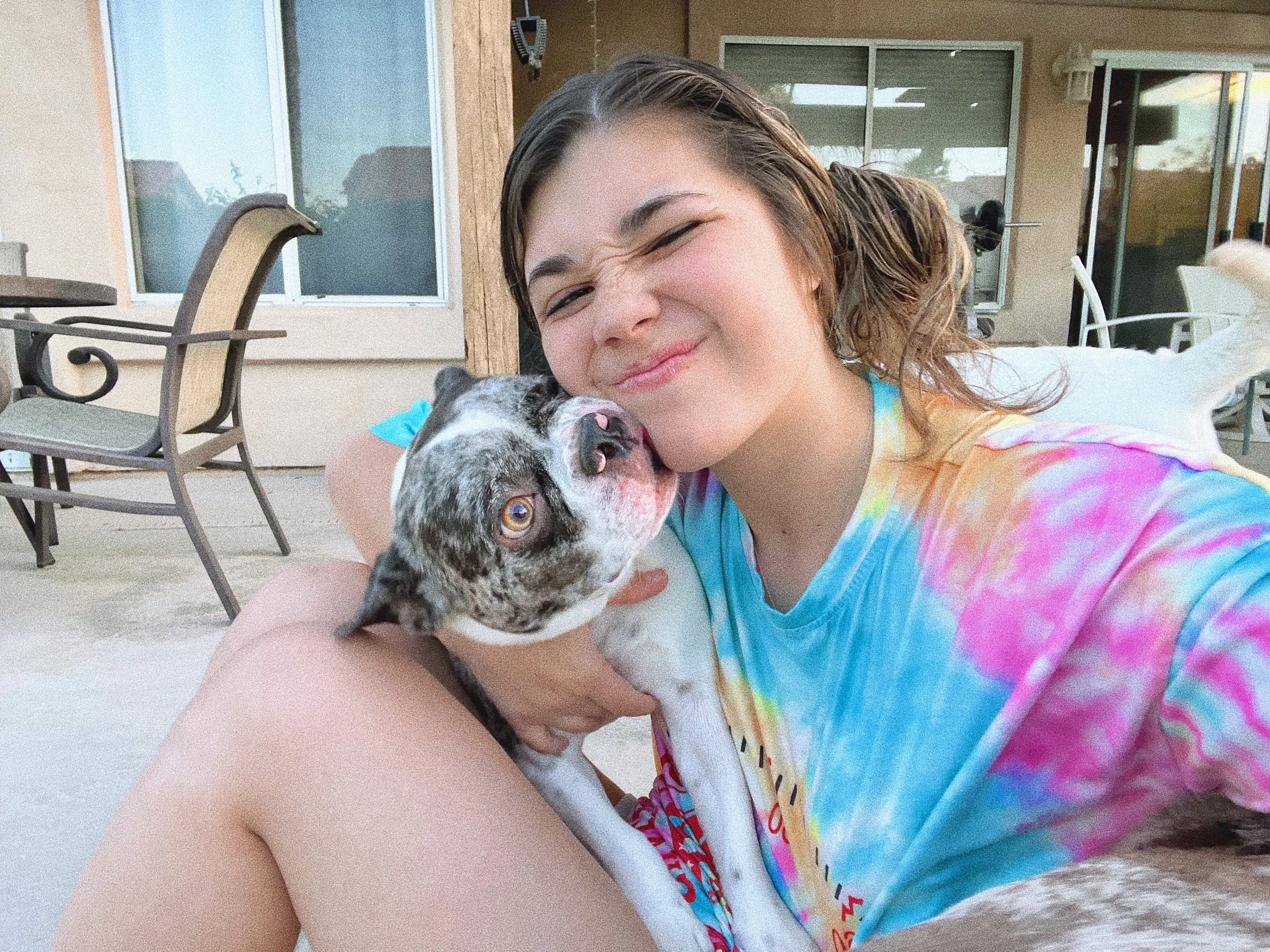 Young woman with wet hair and colorful tie-dye shirt cuddling a small brindle and white dog outdoors on a patio, smiling and squinting at the camera-Dog Board & Train in La Quinta CA