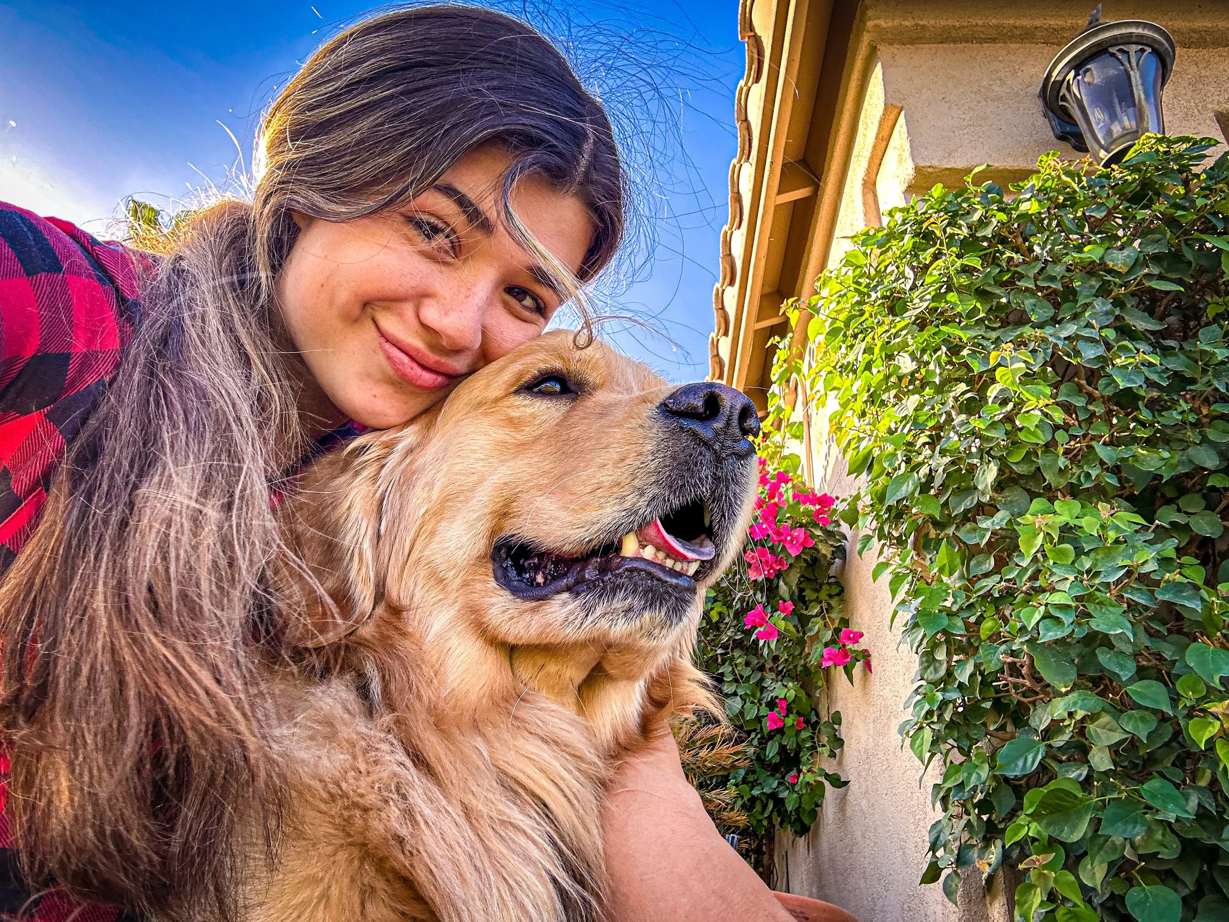A young woman taking a selfie outdoors with her golden retriever dog, both smiling, next to a leafy green bush with pink flowers, and a beige house with a lantern-Dog Board & Train in La Quinta CA