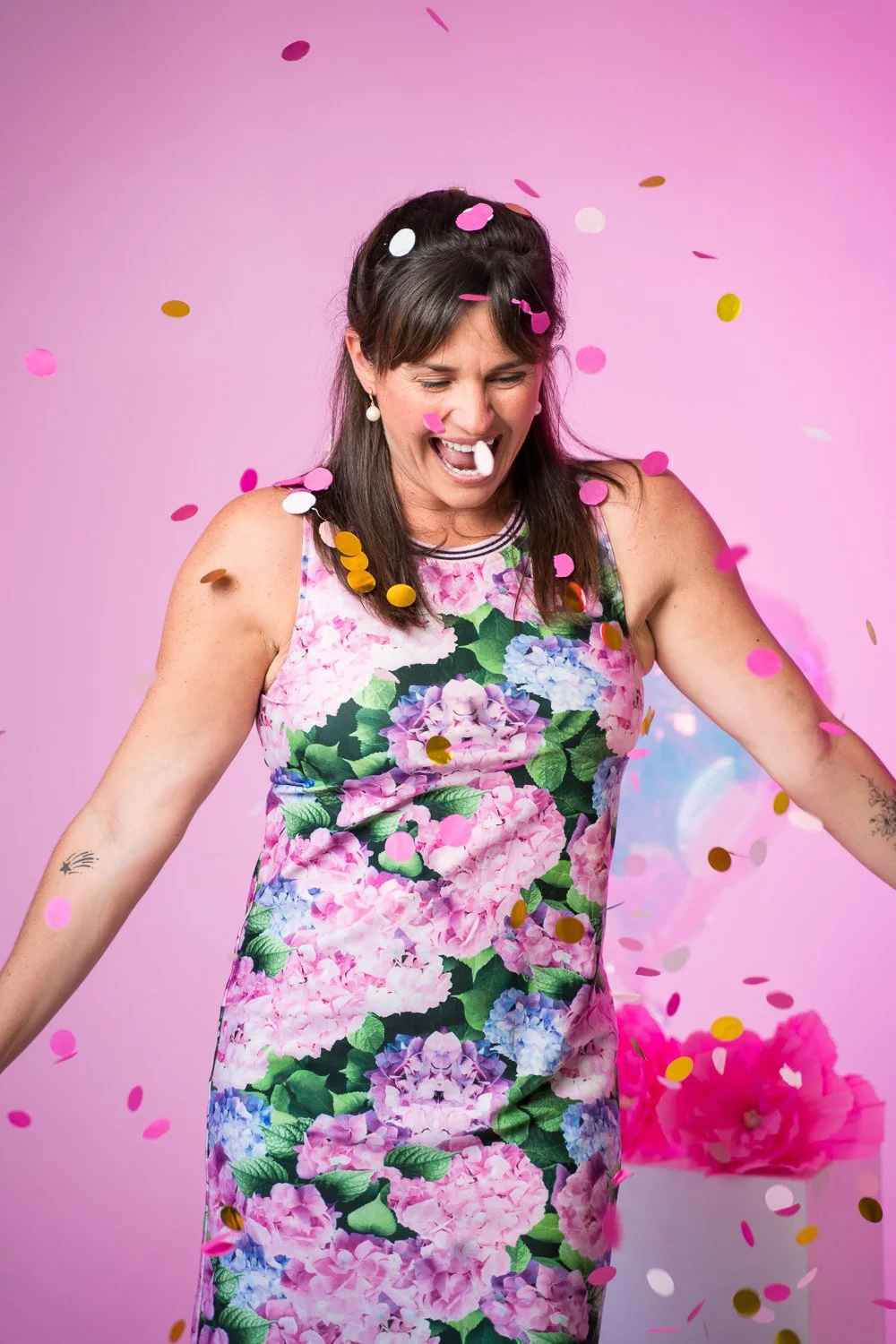 Woman celebrating with confetti, wearing a floral dress, against a pink background.