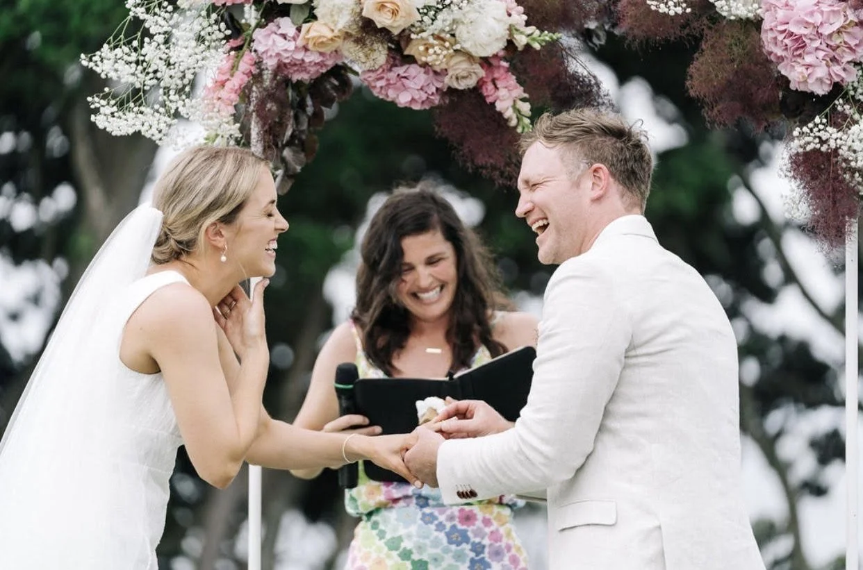 A bride and groom exchange wedding vows at an outdoor wedding ceremony, smiling and holding hands, with an officiant standing behind them under a floral arch.
