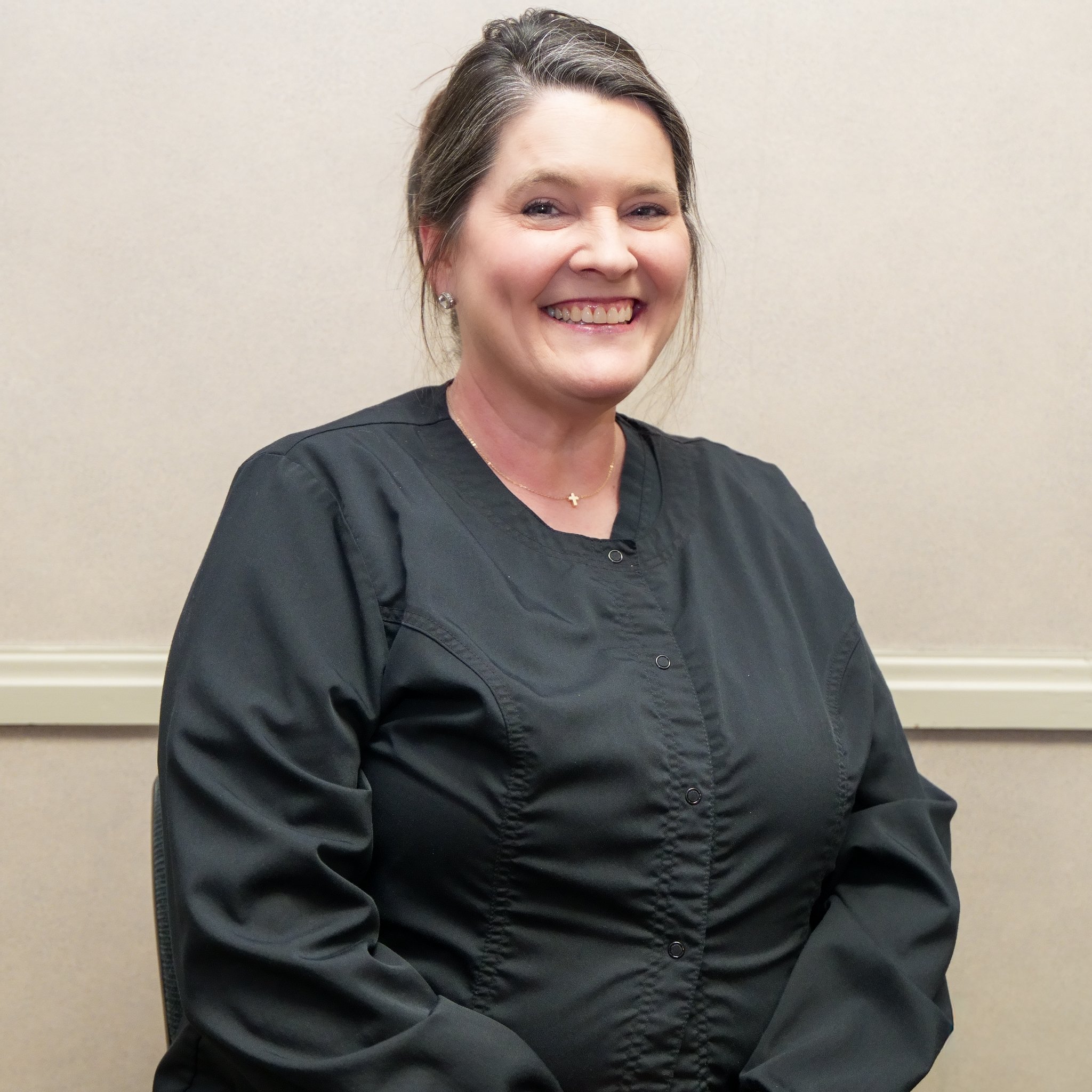 A woman in a black medical uniform smiling, sitting against a beige wall.