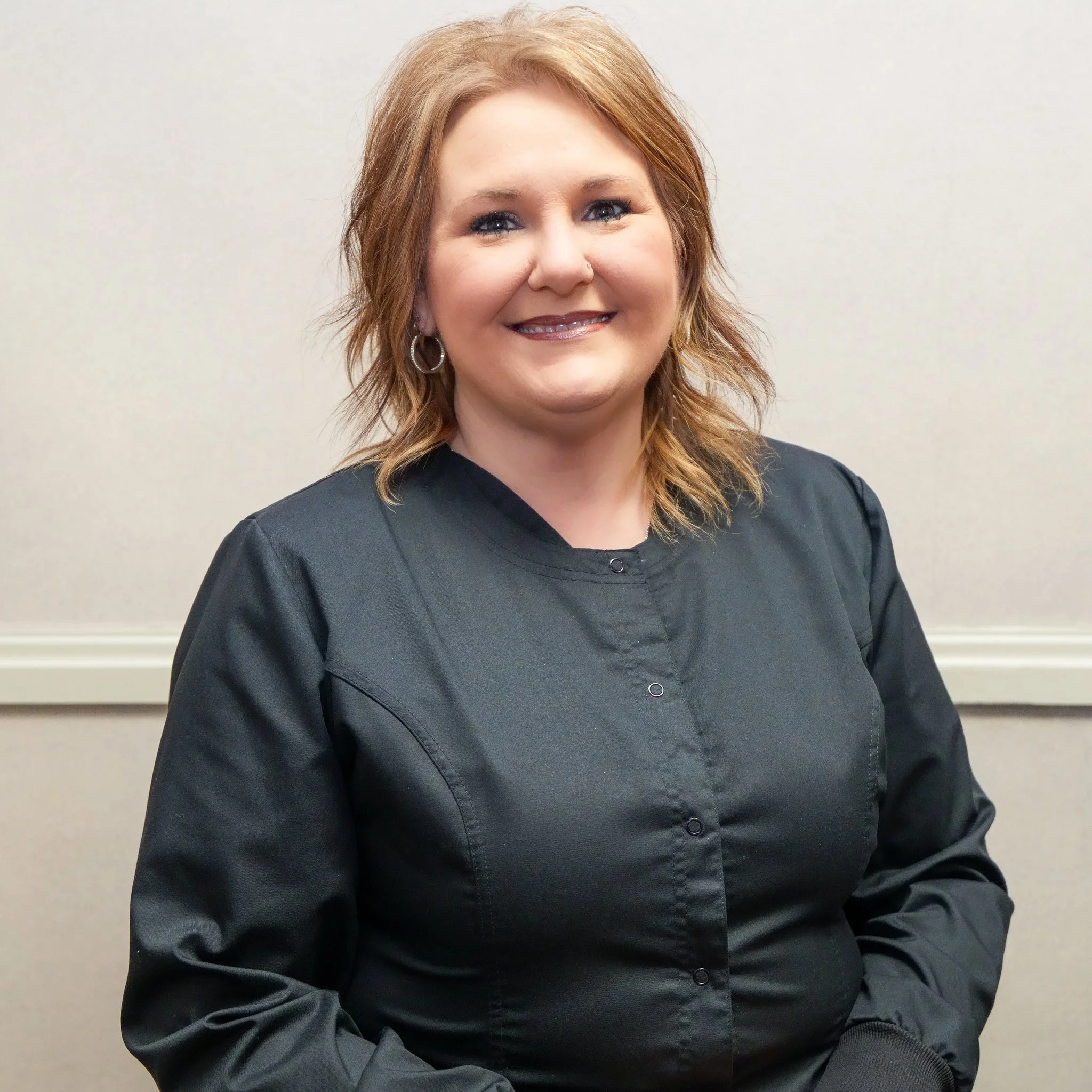A woman with shoulder-length reddish-brown hair, wearing hoop earrings and a black button-up shirt, smiling against a plain beige wall.