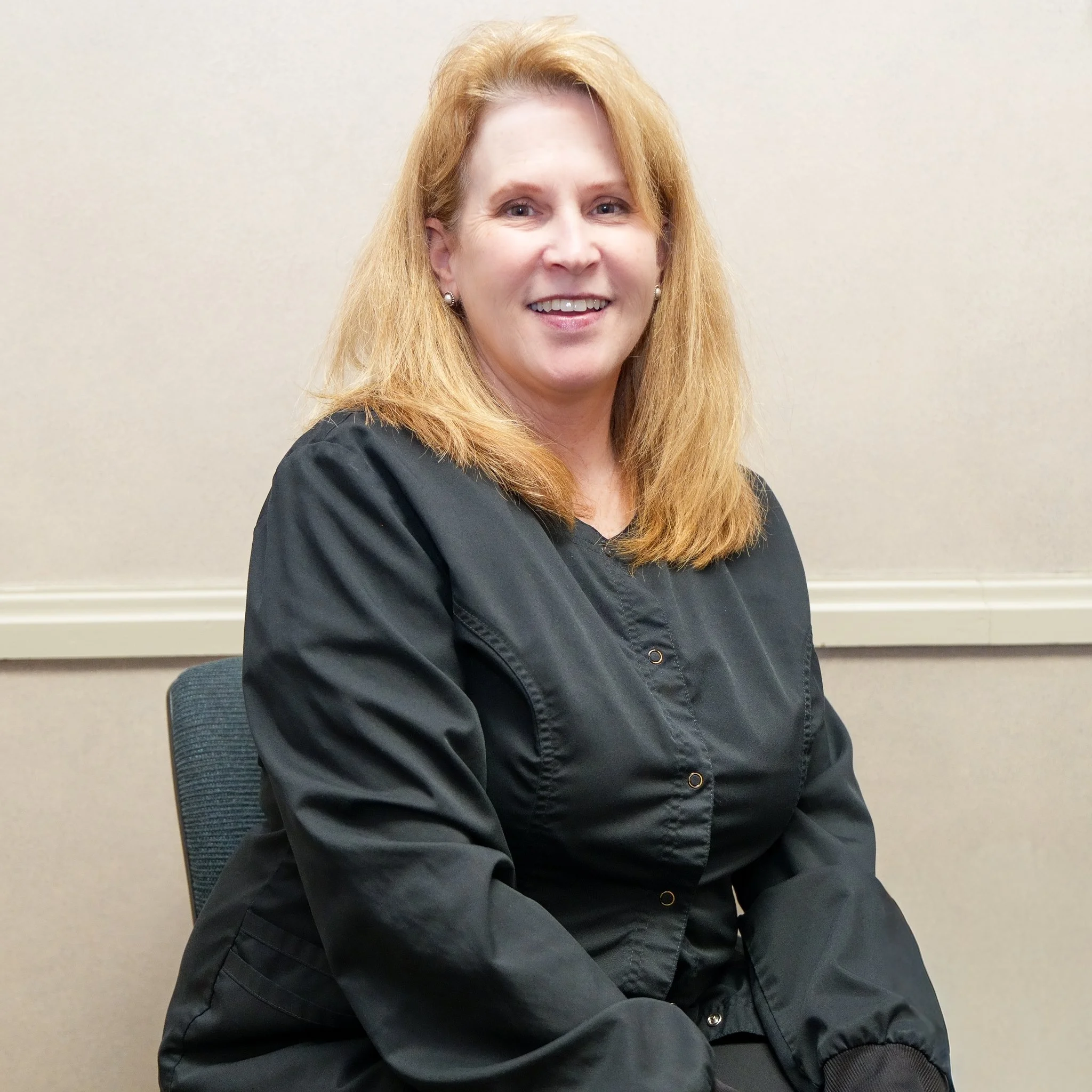 A woman with shoulder-length red hair, wearing a black jacket, seated and smiling in front of a plain beige wall.