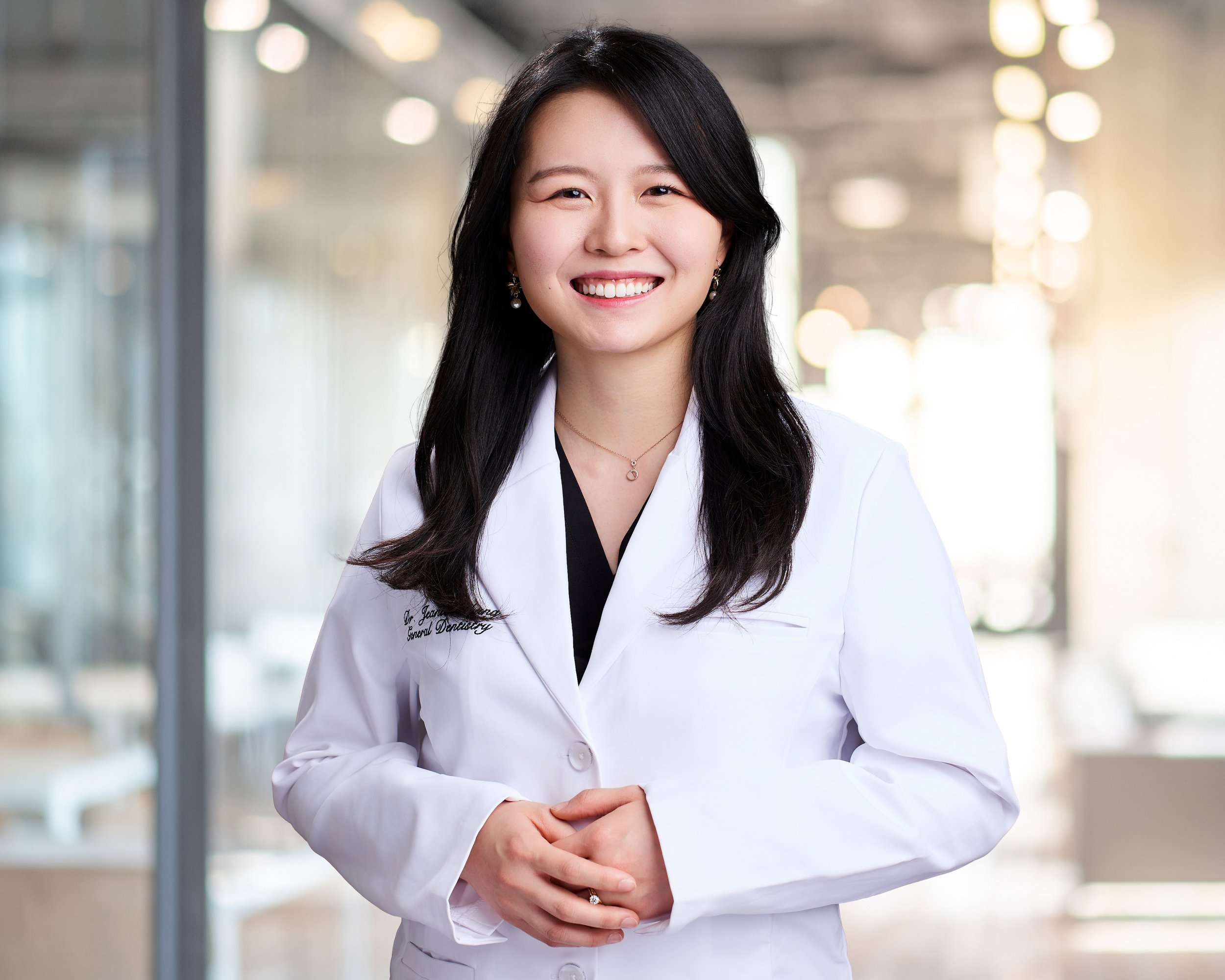Smiling female doctor wearing a white coat with her hands clasped in front of her in a bright, modern medical office.