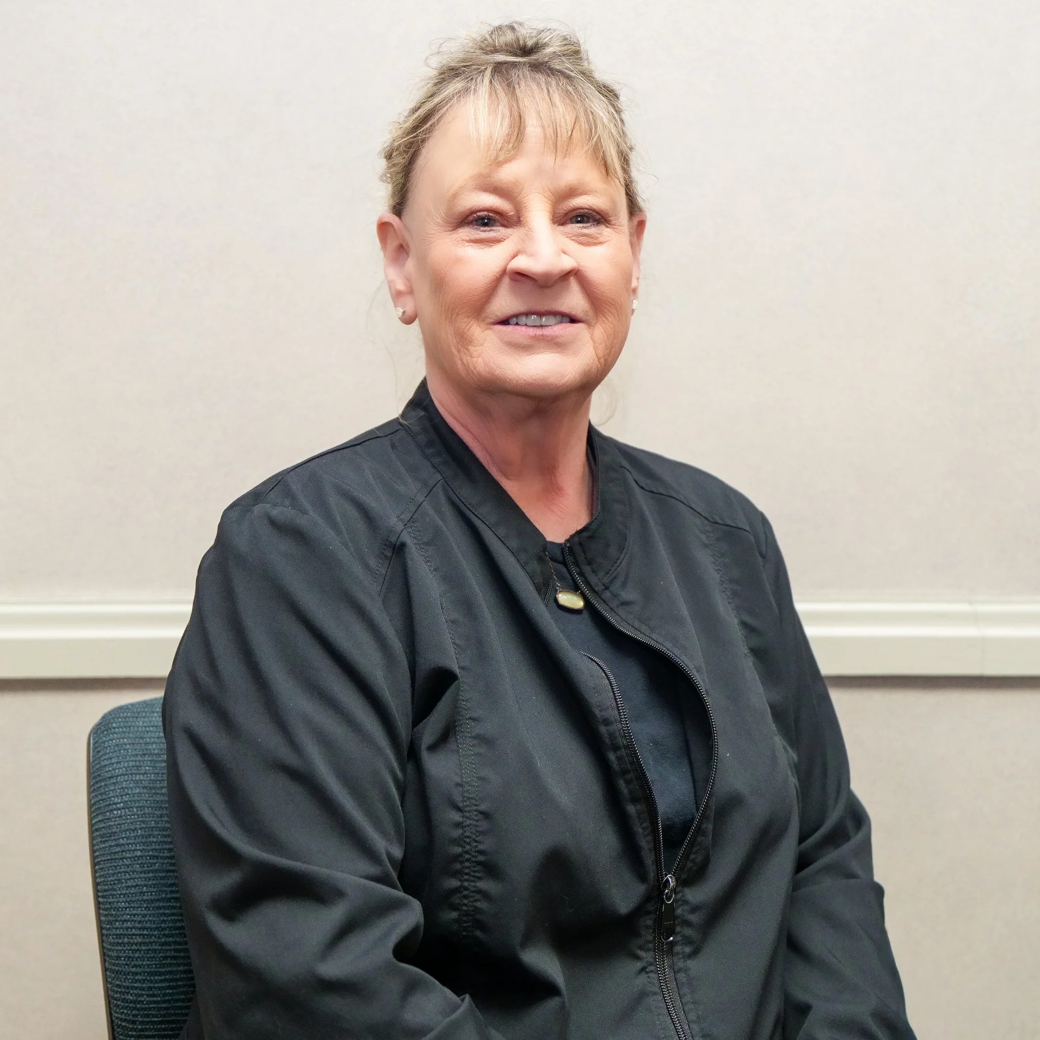 A middle-aged woman with blonde hair in a casual black jacket, sitting against a beige wall with a chair.