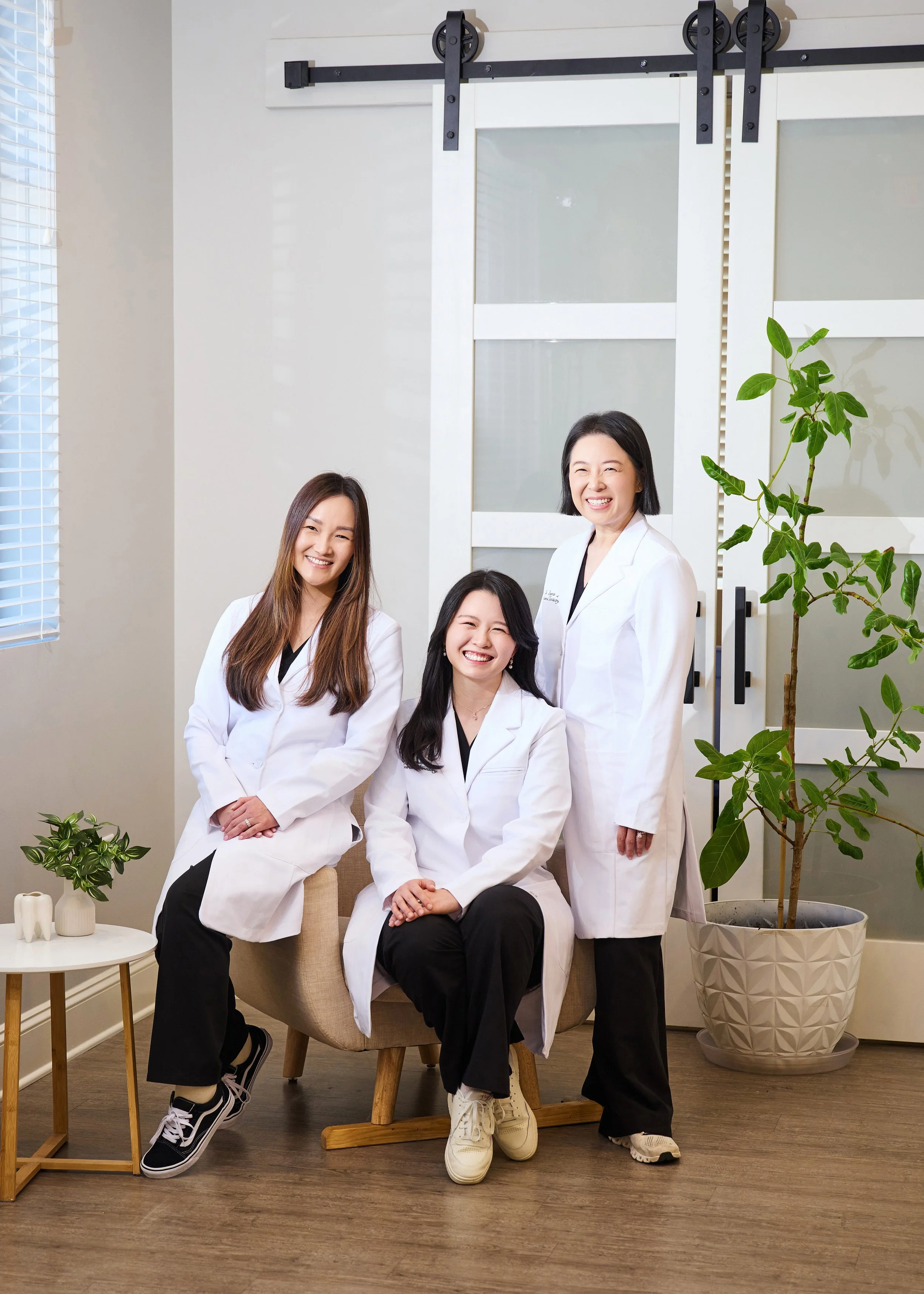 Three women in white lab coats smiling in a modern medical or office setting with plants and wooden furniture.