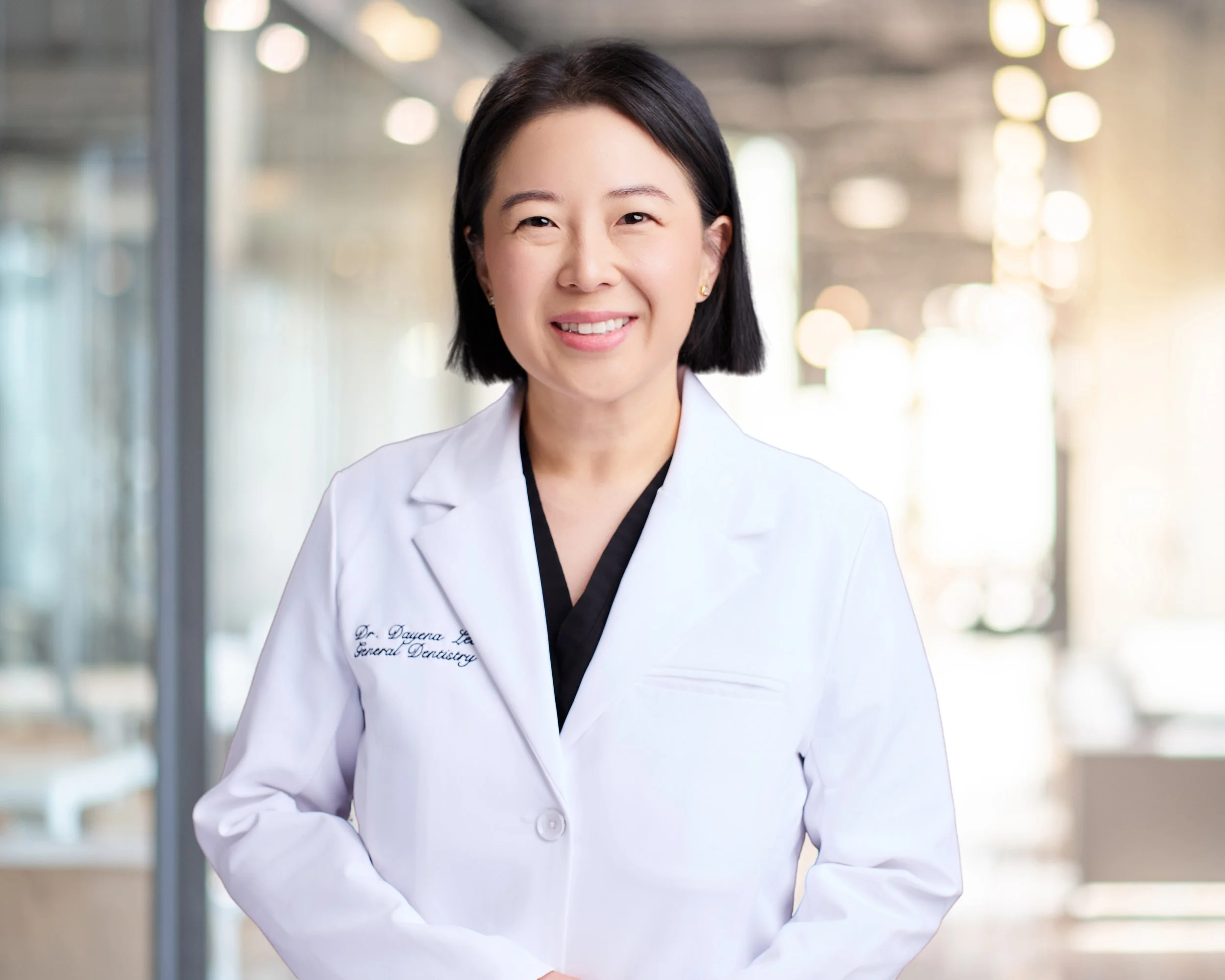 A woman with black hair in a bob haircut, smiling, wearing a white professional coat with embroidered text, in a bright modern office or dental clinic setting.