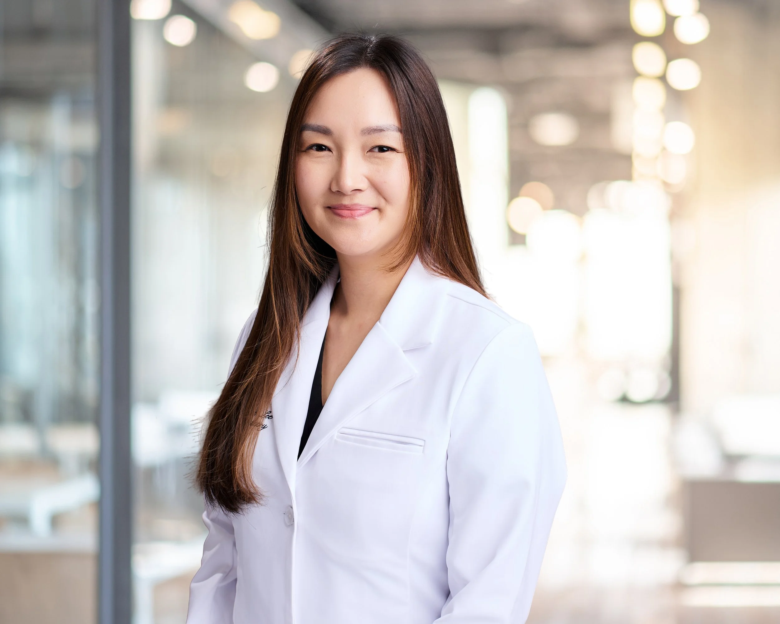 Portrait of a female medical professional wearing a white coat, smiling in a bright clinic or hospital corridor.