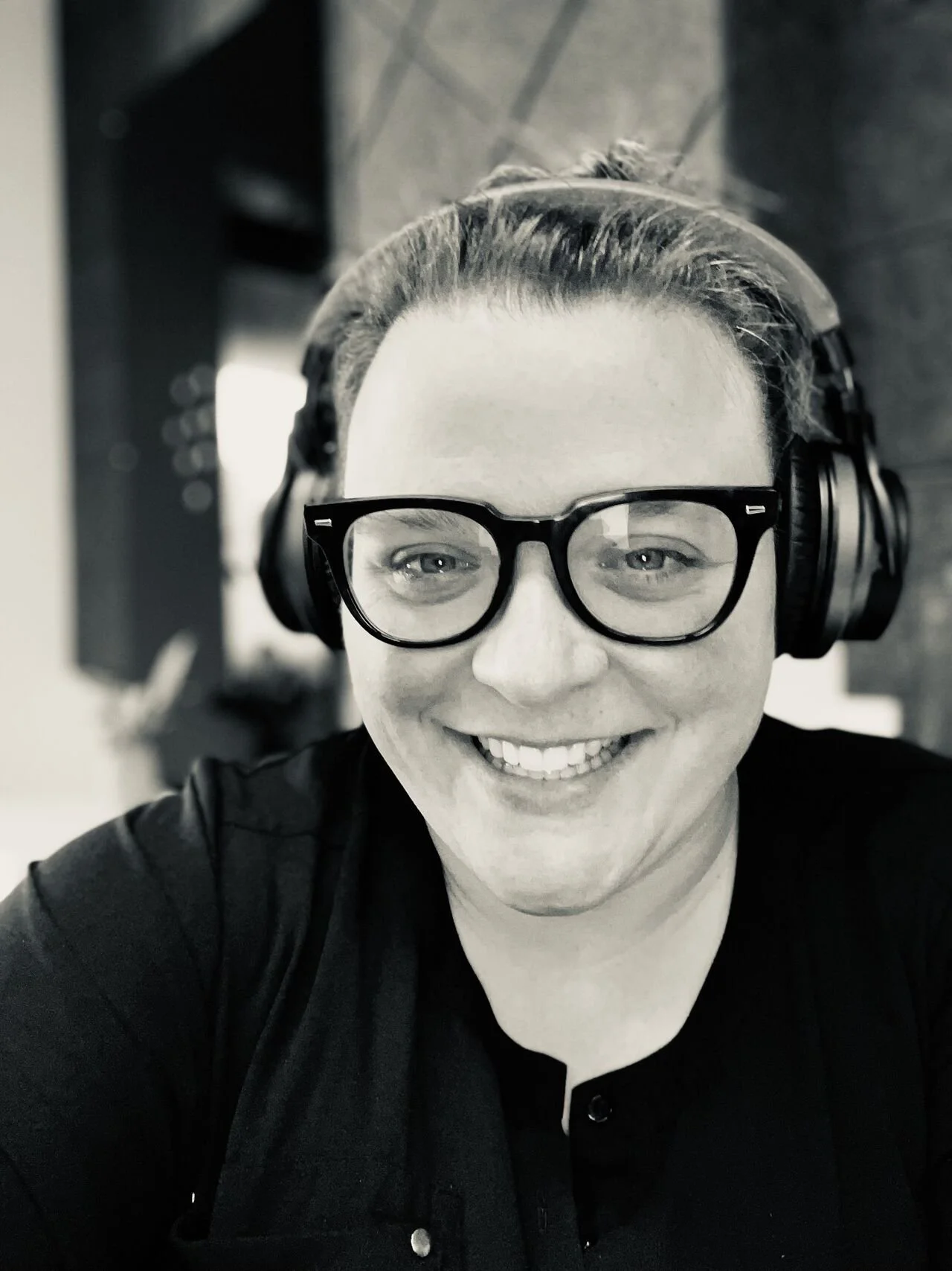 A person smiling, wearing glasses and headphones, with dark hair pulled back, in black and white photo.