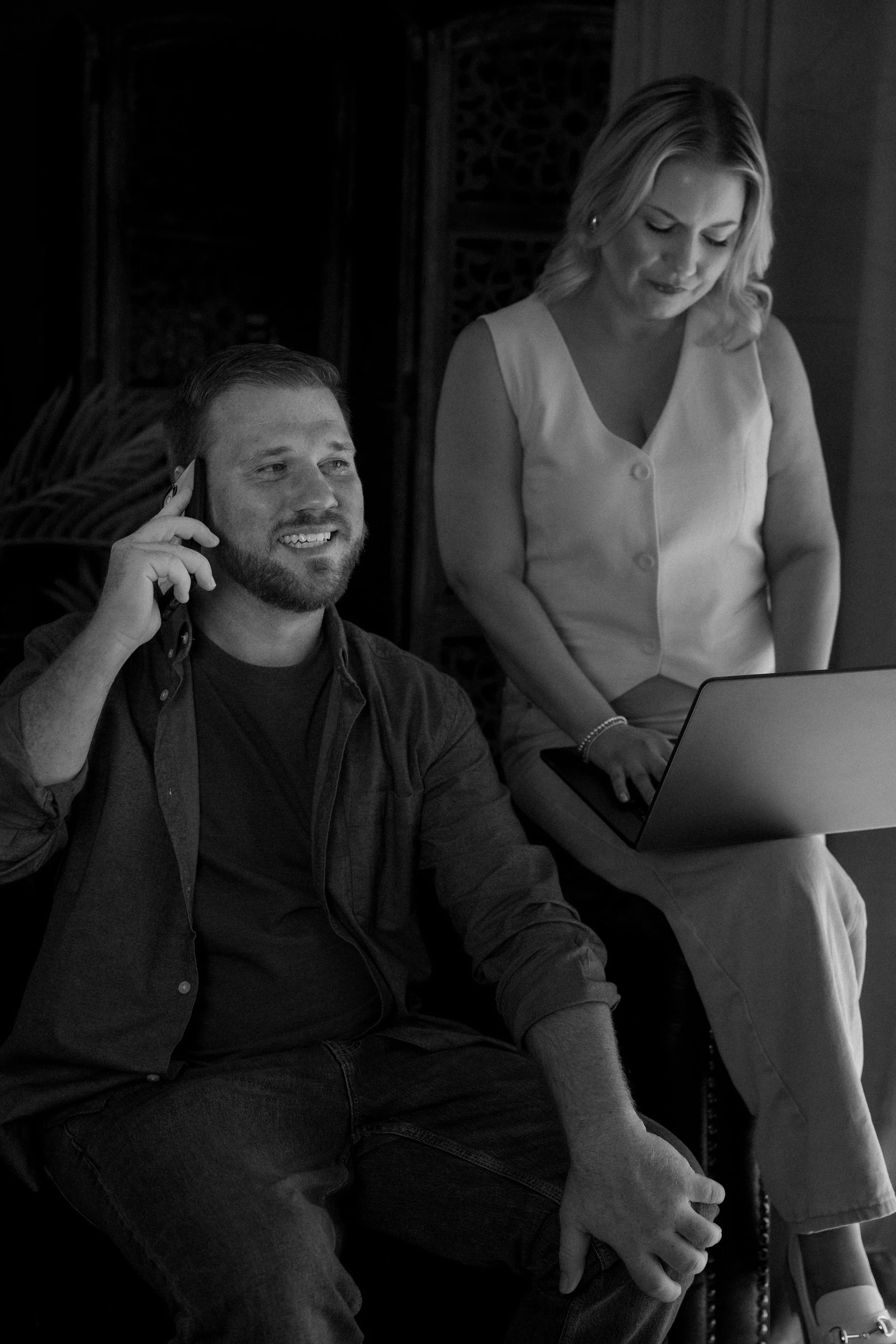 A black and white photo of a man sitting on a chair, talking on the phone, with a woman standing beside him looking down at a laptop computer.