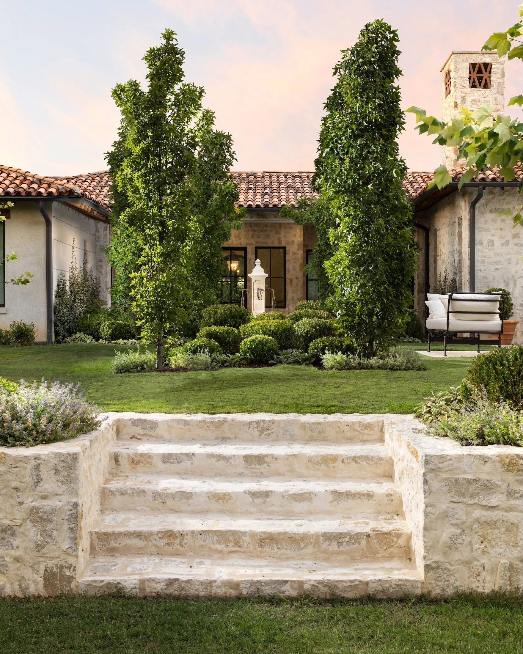 Stone steps leading up to a house with lush green trees and bushes in the garden. The house has a tiled roof, a window with black trim, and a stone chimney. There is outdoor furniture on the patio.
