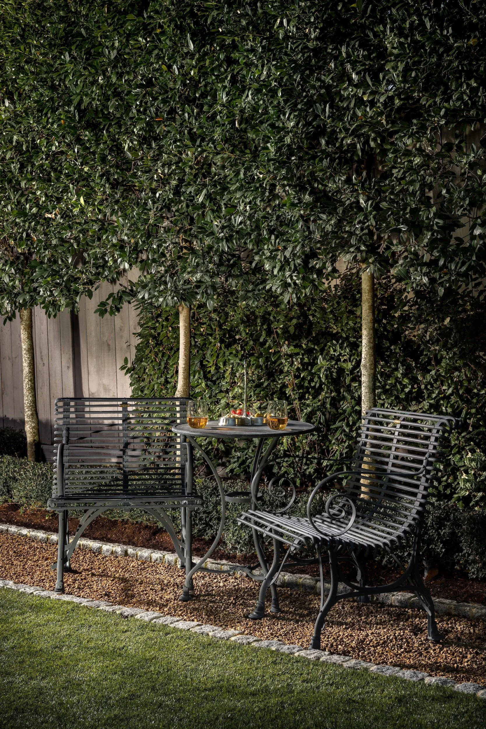 Nighttime outdoor patio with metal chairs, a small round table with drinks, a deer figurine, and a lush green hedge background.