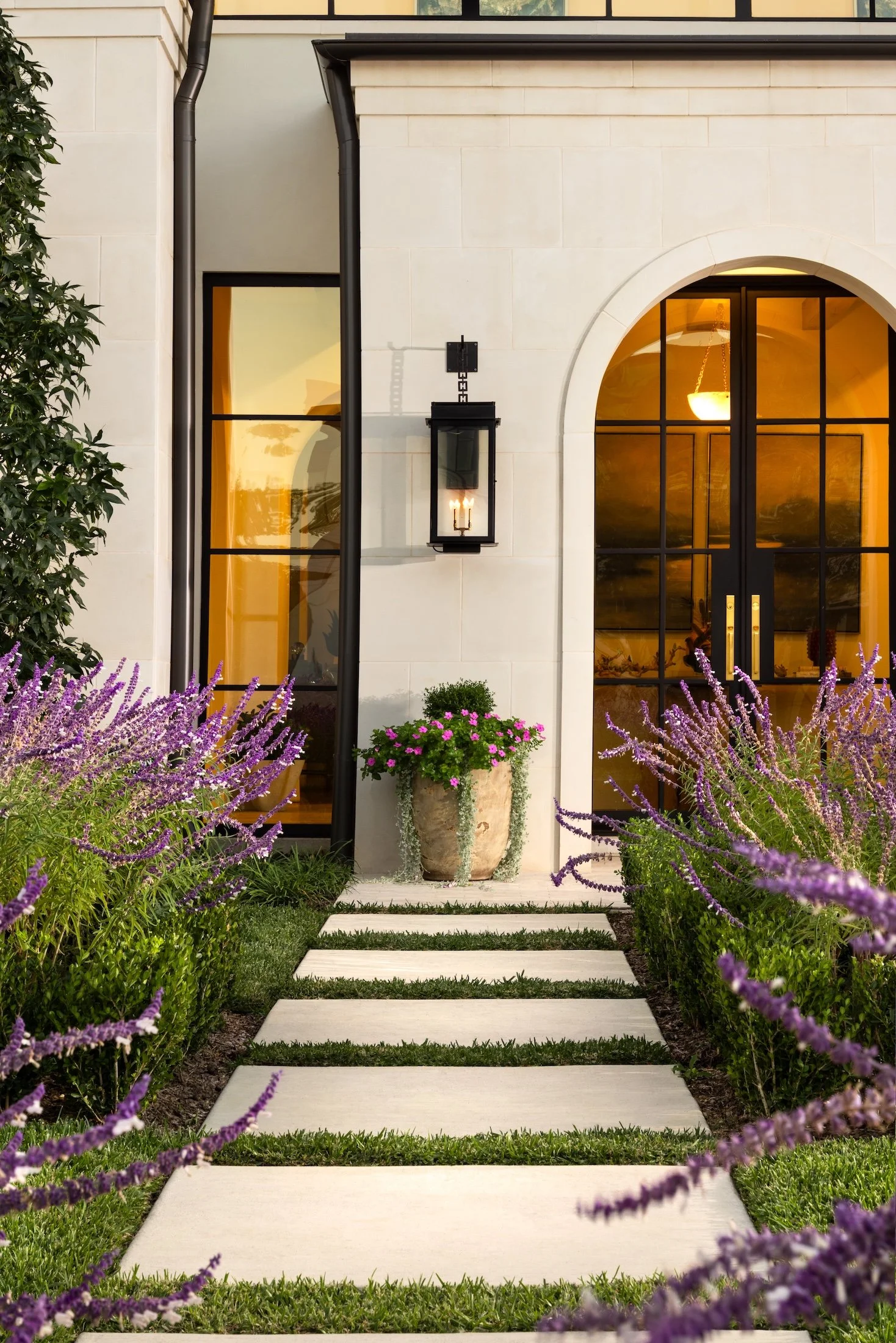 Front view of a house with large arched window and glass door, surrounded by lavender flowers and a stone pathway leading to the entrance, with a lantern-style light fixture on the wall.