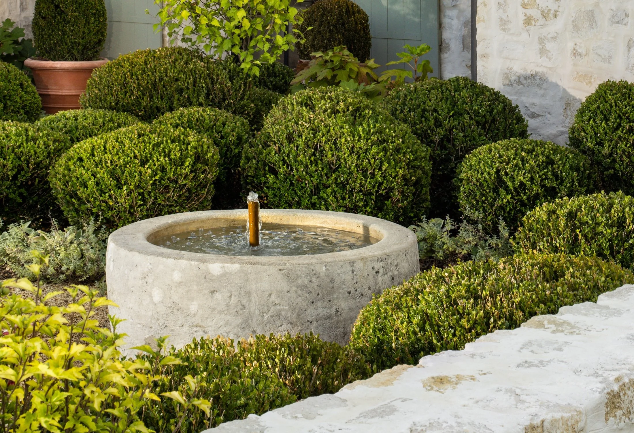 A garden with a round concrete fountain with a bubbling water spout, surrounded by neatly trimmed bushes, plants, and a stone wall with a blue door in the background.
