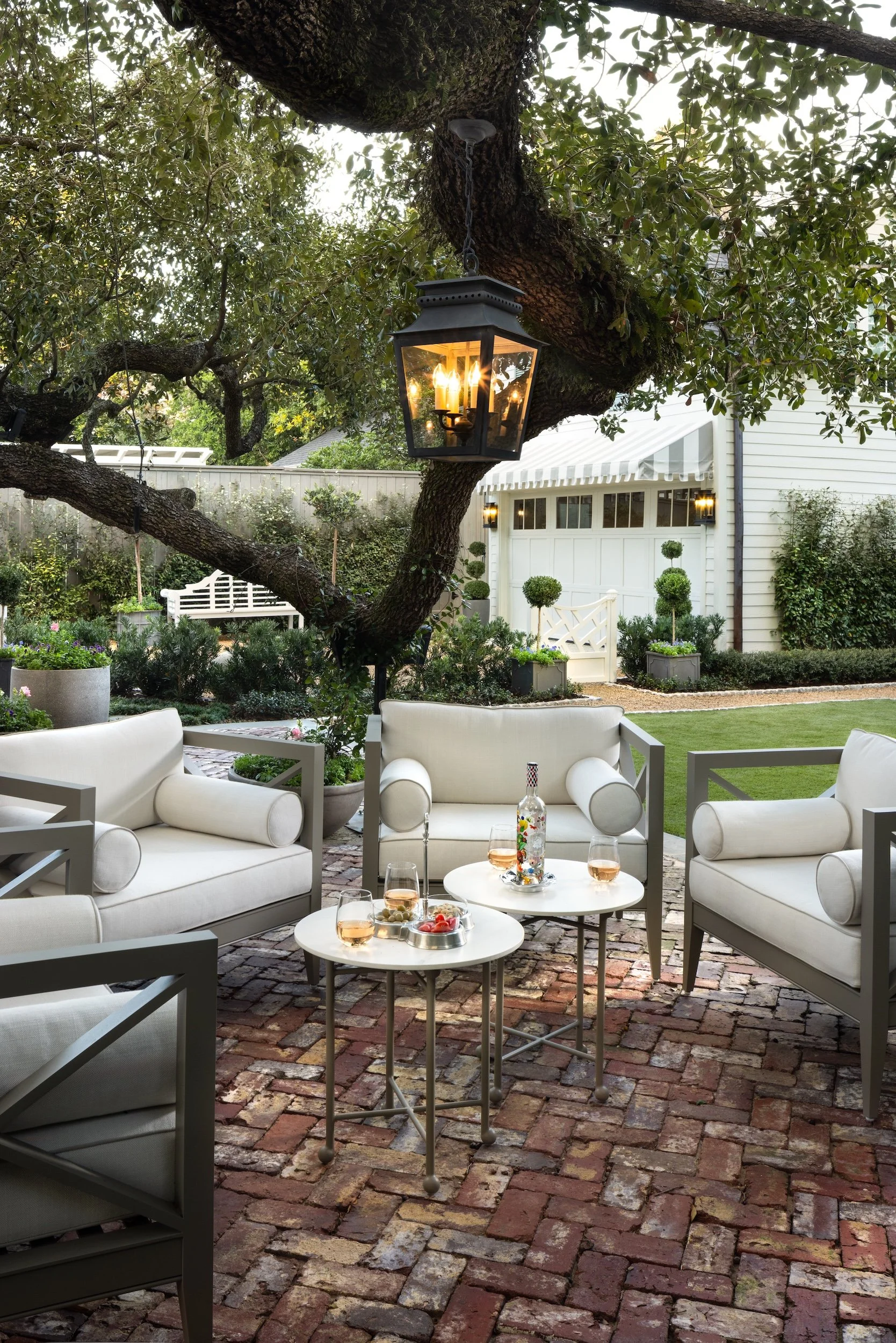 A cozy outdoor patio area with white seating and a small round white table set with glasses of rosé wine and snacks. A brick floor and lush greenery surround the space, with a large tree overhead and a lantern hanging from its branch. In the background, a white house with a decorated front yard and outdoor lights.