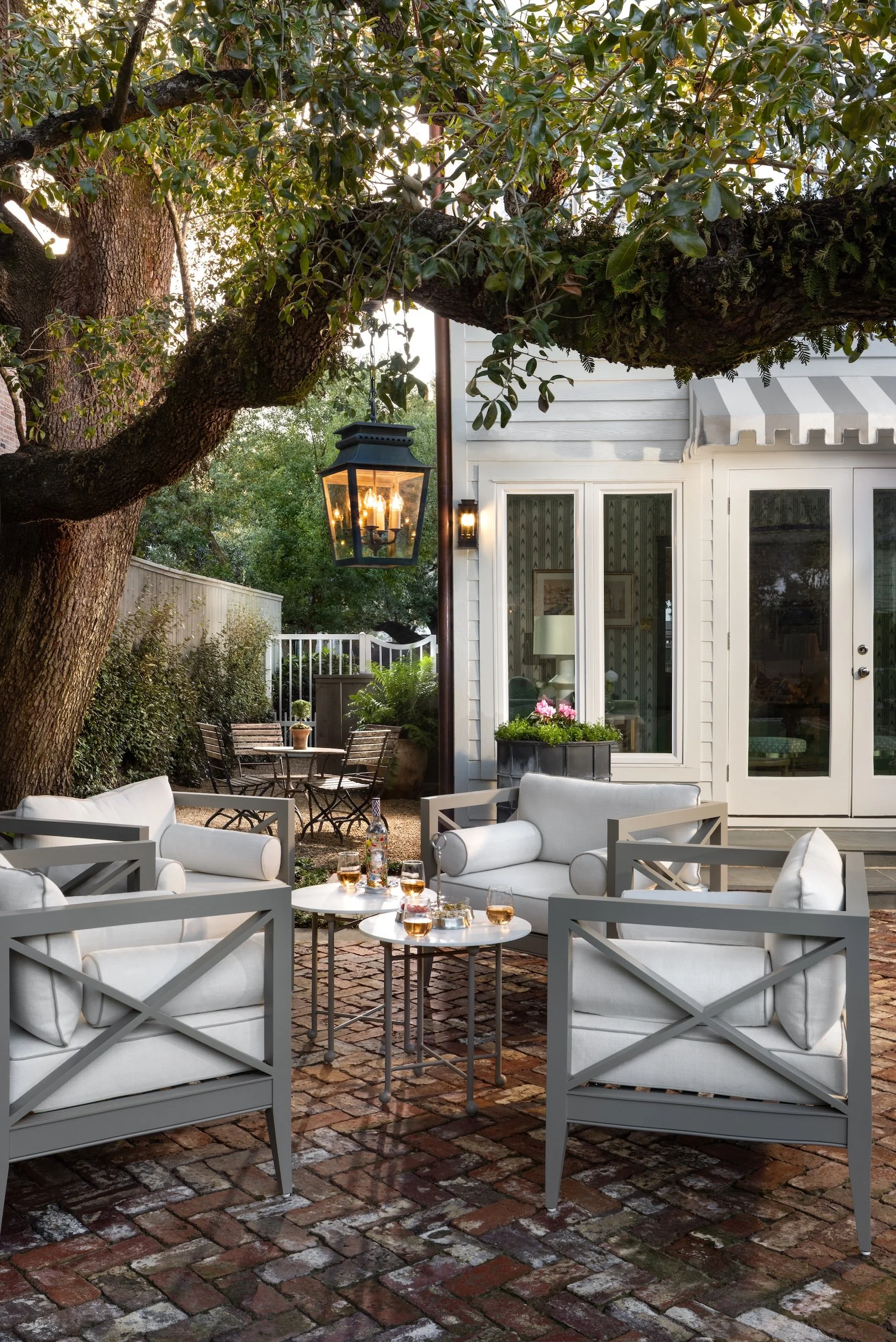 An outdoor patio area with white cushioned seating, small round tables with drinks, a large tree with dense foliage, and a house with large windows and a striped awning, illuminated by outdoor lanterns and surrounded by greenery.