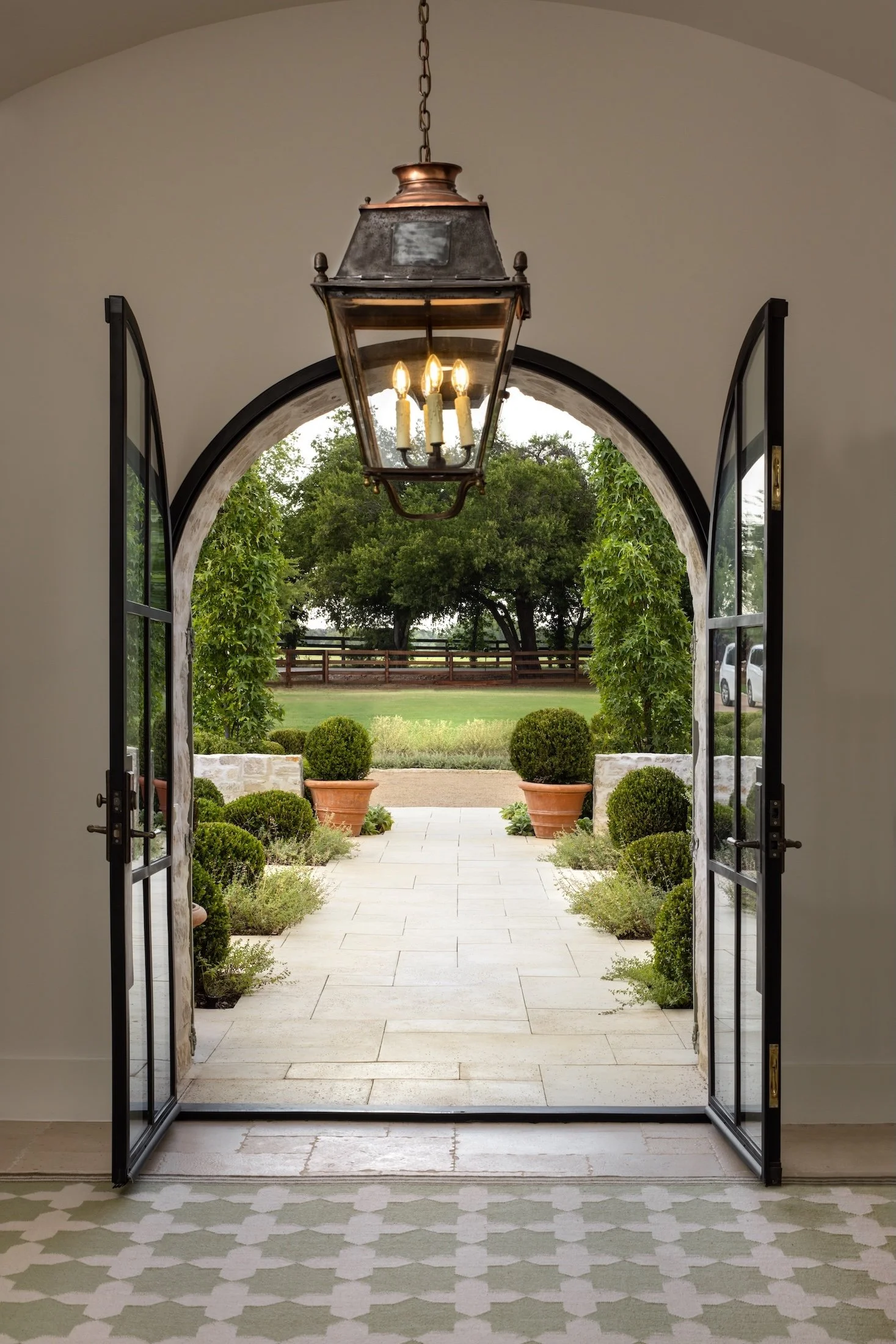 Open glass double doors leading to a courtyard with potted bushes, trees, a wooden fence, and parked cars in the background, with a vintage chandelier hanging from the ceiling.