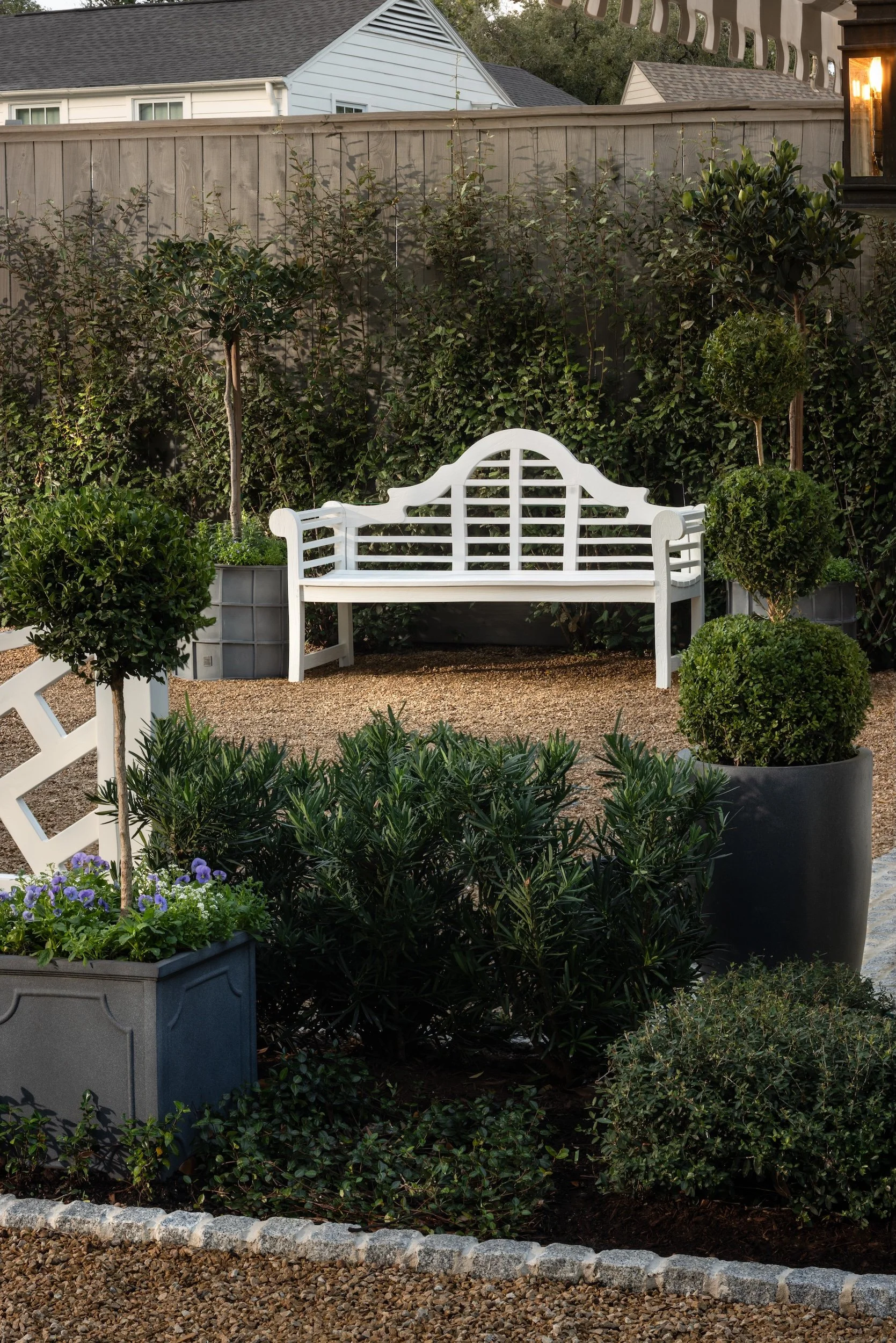 A backyard garden with a white decorative bench, various bushes, potted plants, and flowering plants, enclosed by a wooden fence, with neighboring houses in the background.