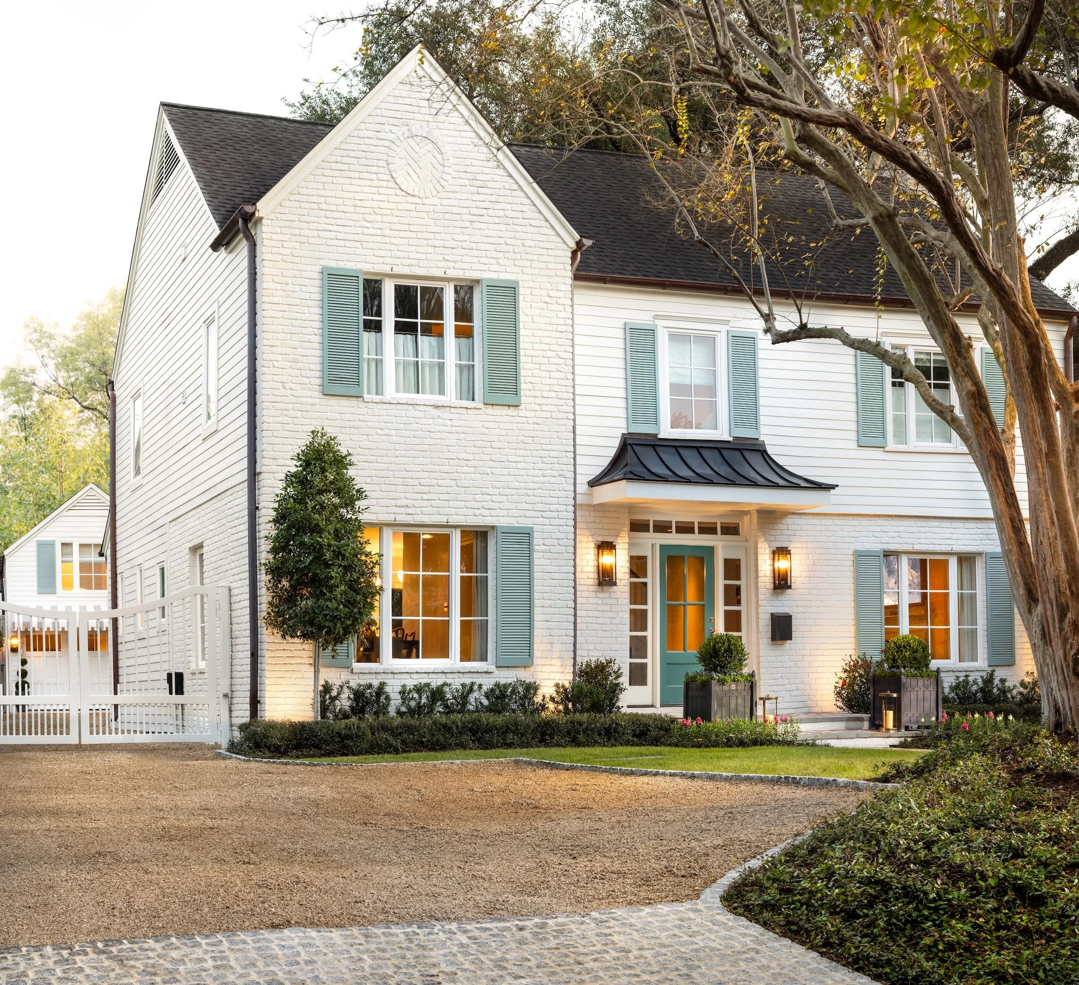 Front view of a white two-story house with a sloped black roof, multiple windows with light blue shutters, a front door with glass panes, and a yard with a gravel driveway, small bushes, and trees.