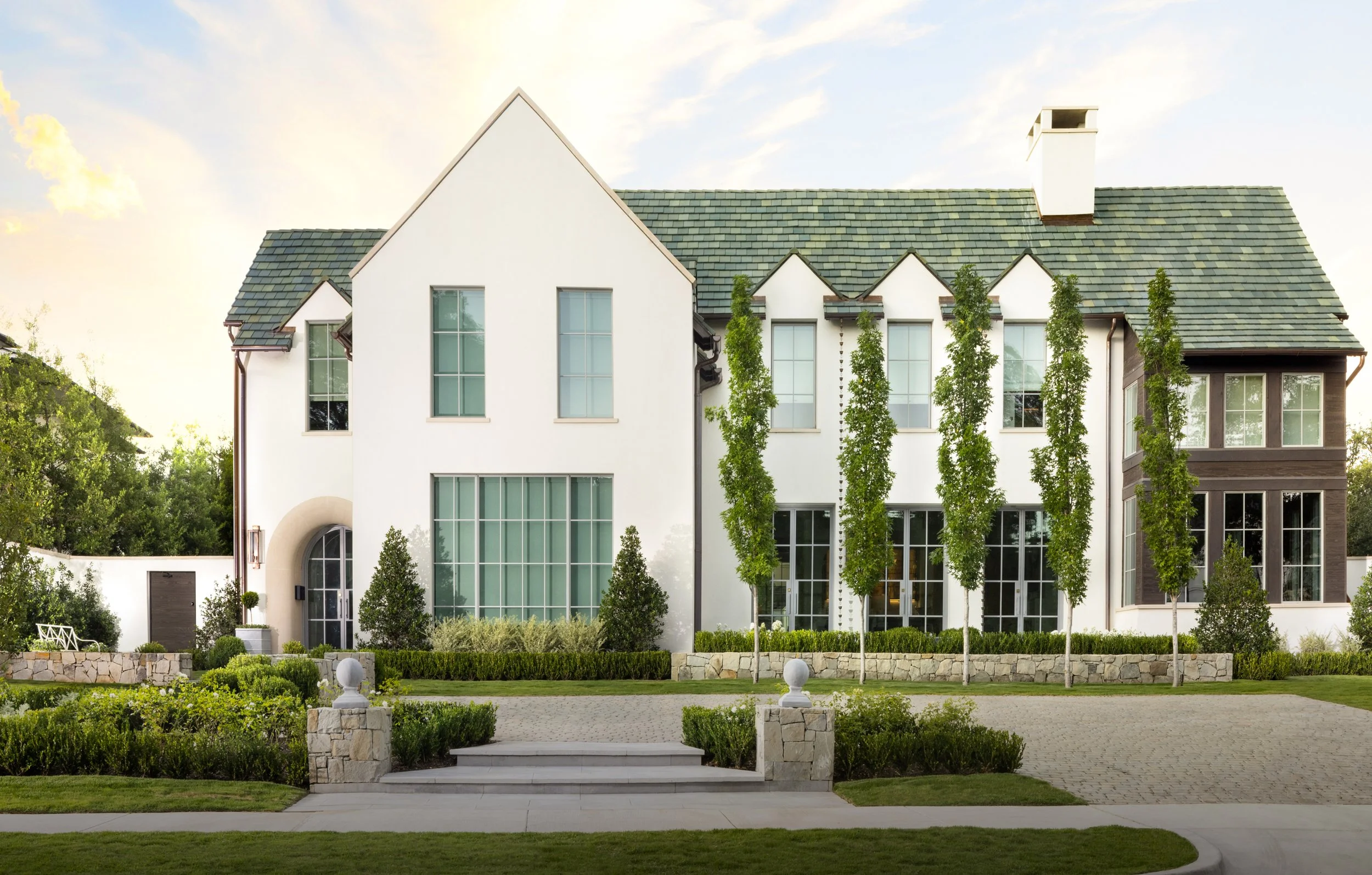 Modern multi-story house with white walls, large glass windows, a green shingle roof, a chimney, and a landscaped front yard with trees and shrubs at sunset.
