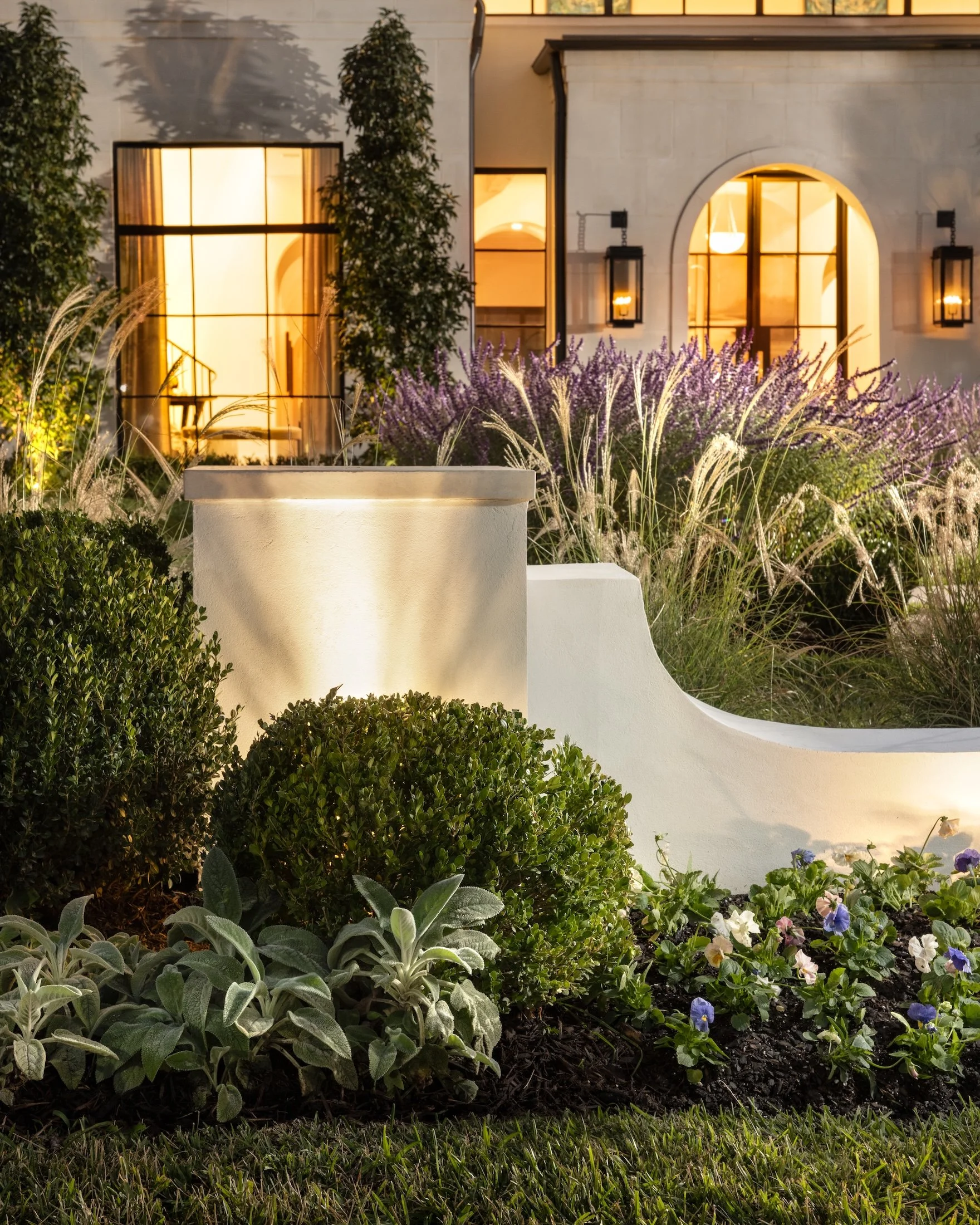 Night view of a well-lit house with large windows, surrounded by lush Greenery and blooming purple and white flowers, with outdoor lighting illuminating the landscape.