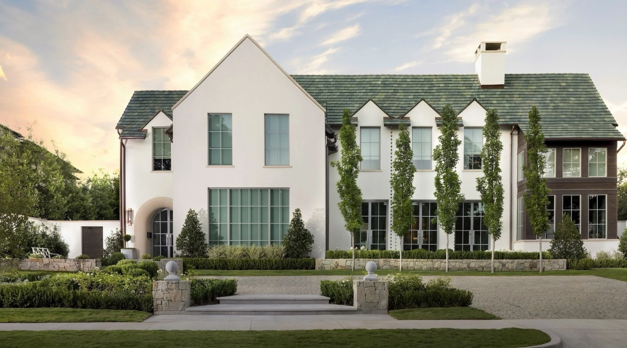 A modern white house with large windows, a green shingle roof, and tall trees in the yard, with a sunset sky in the background.