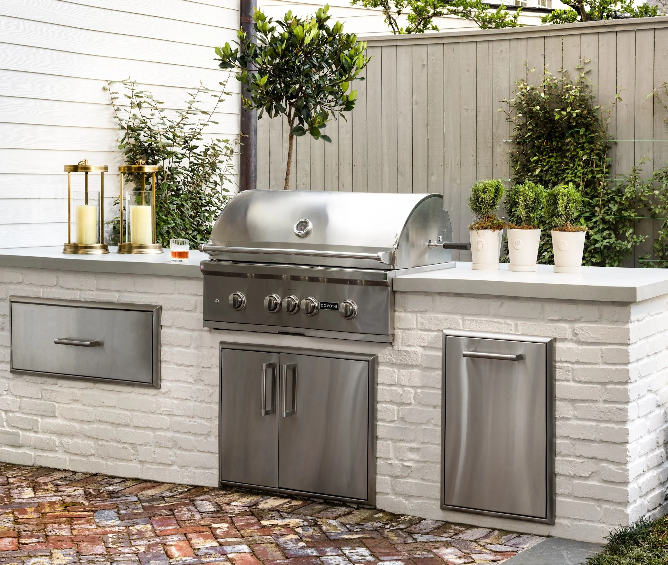 Outdoor kitchen with a stainless steel grill, decorative candles in gold lanterns, potted plants, and a wooden fence with greenery.